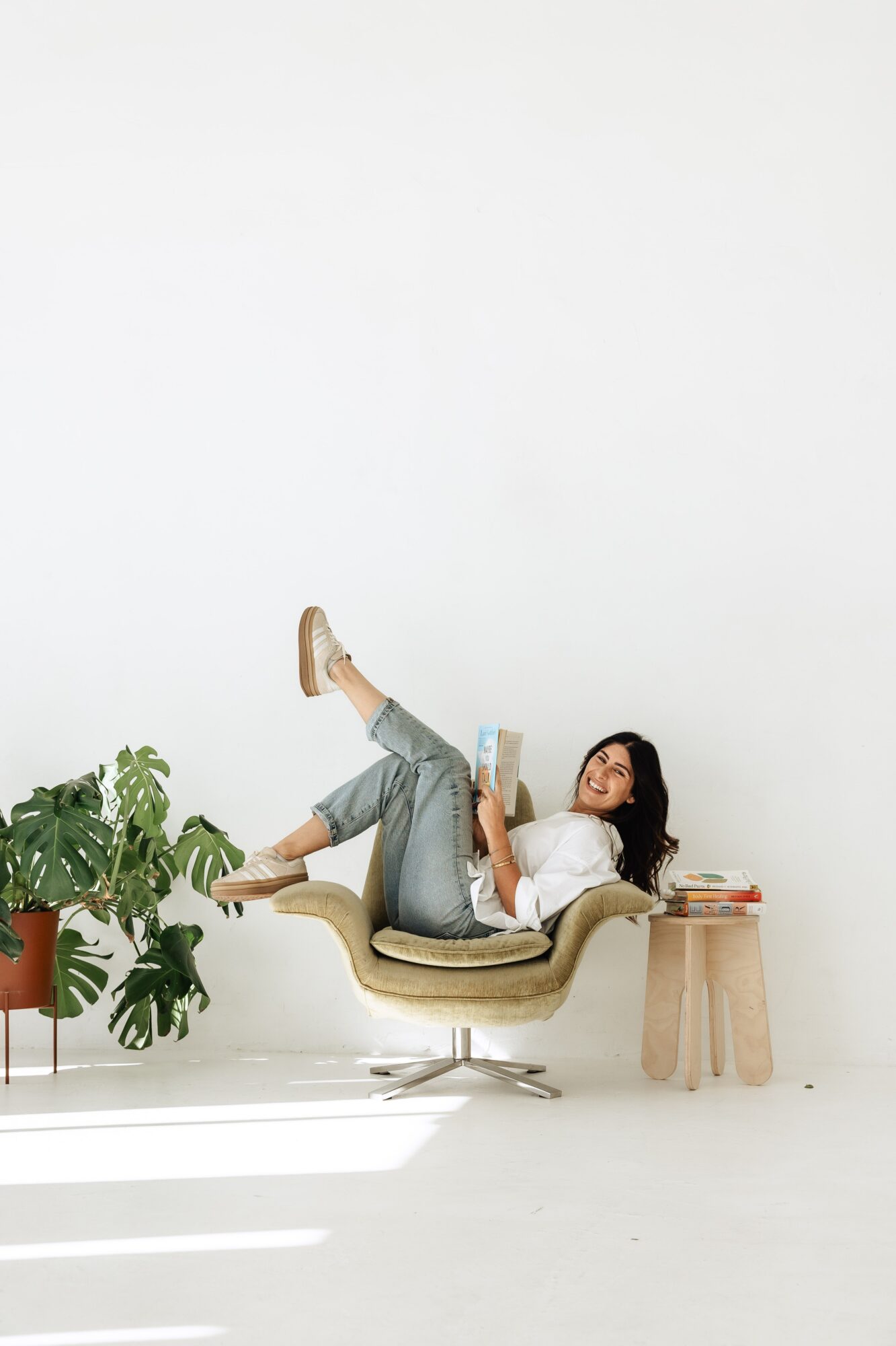 Person relaxing on a chair with a plant and a small table nearby, reading a book, in a bright room.
