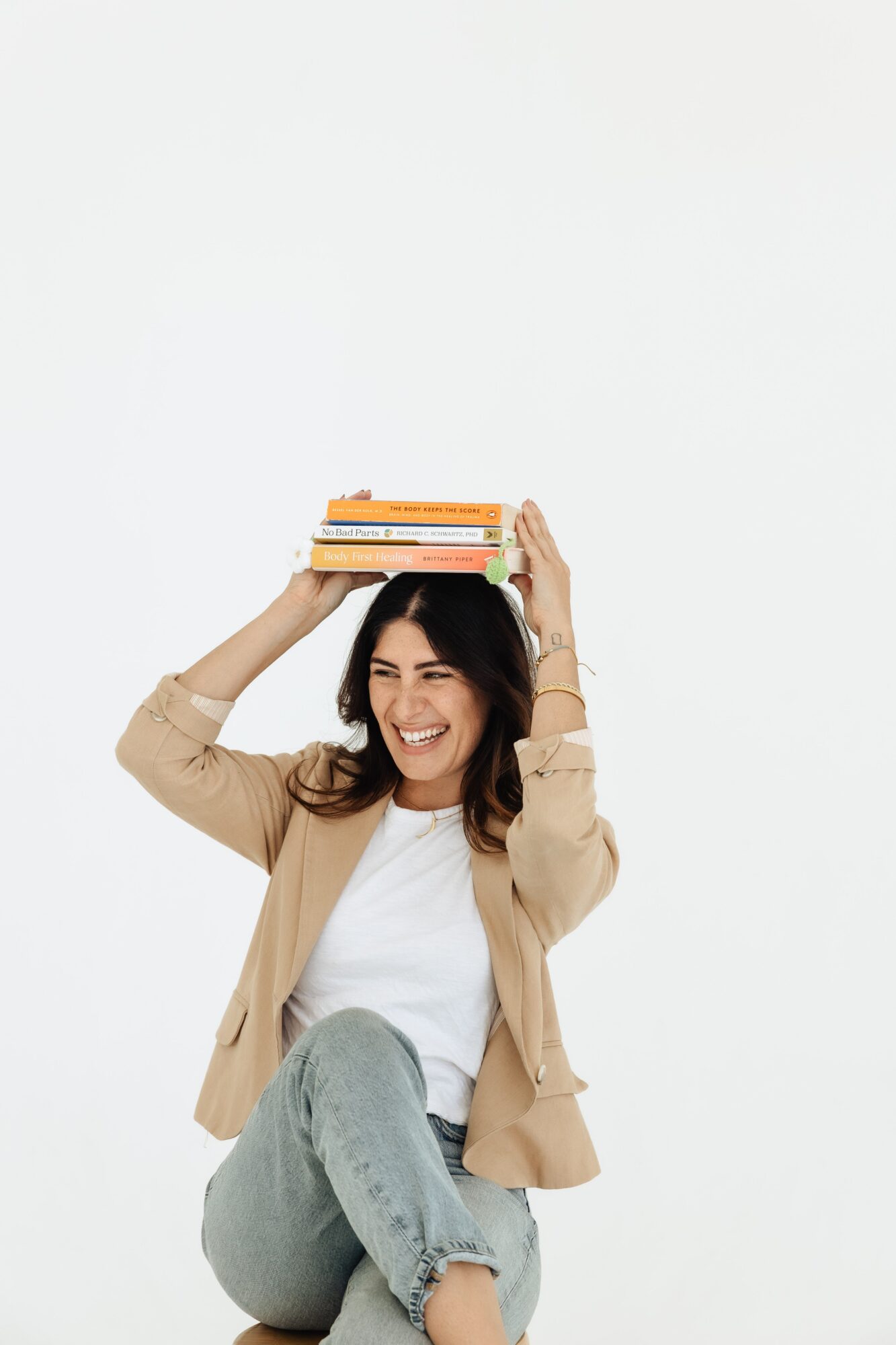 Woman smiling, holding books above her head, sitting cross-legged, wearing a beige jacket and jeans, against a white background.