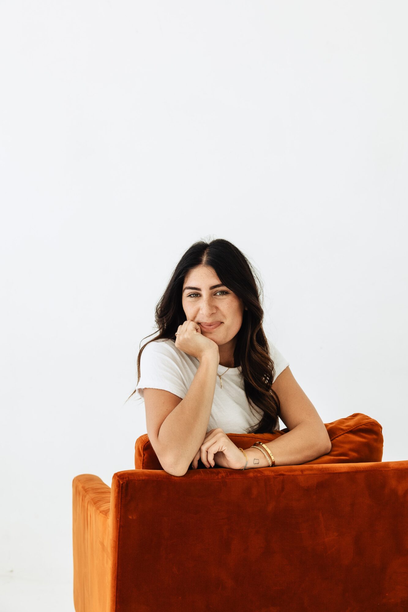 Young woman with dark hair sitting on a brown sofa, resting her chin on her hand, smiling gently, against a plain white background.