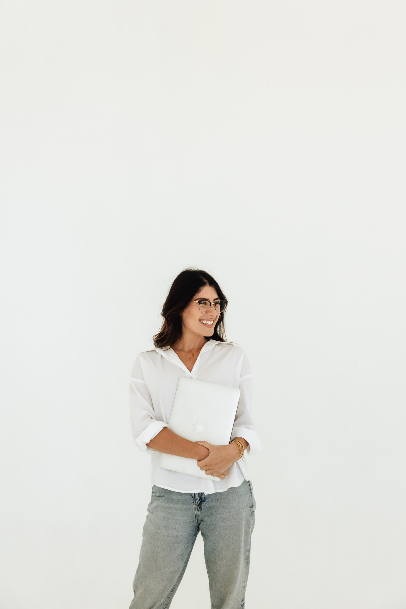 Woman with glasses holding a laptop and papers, smiling, standing against a plain white background.