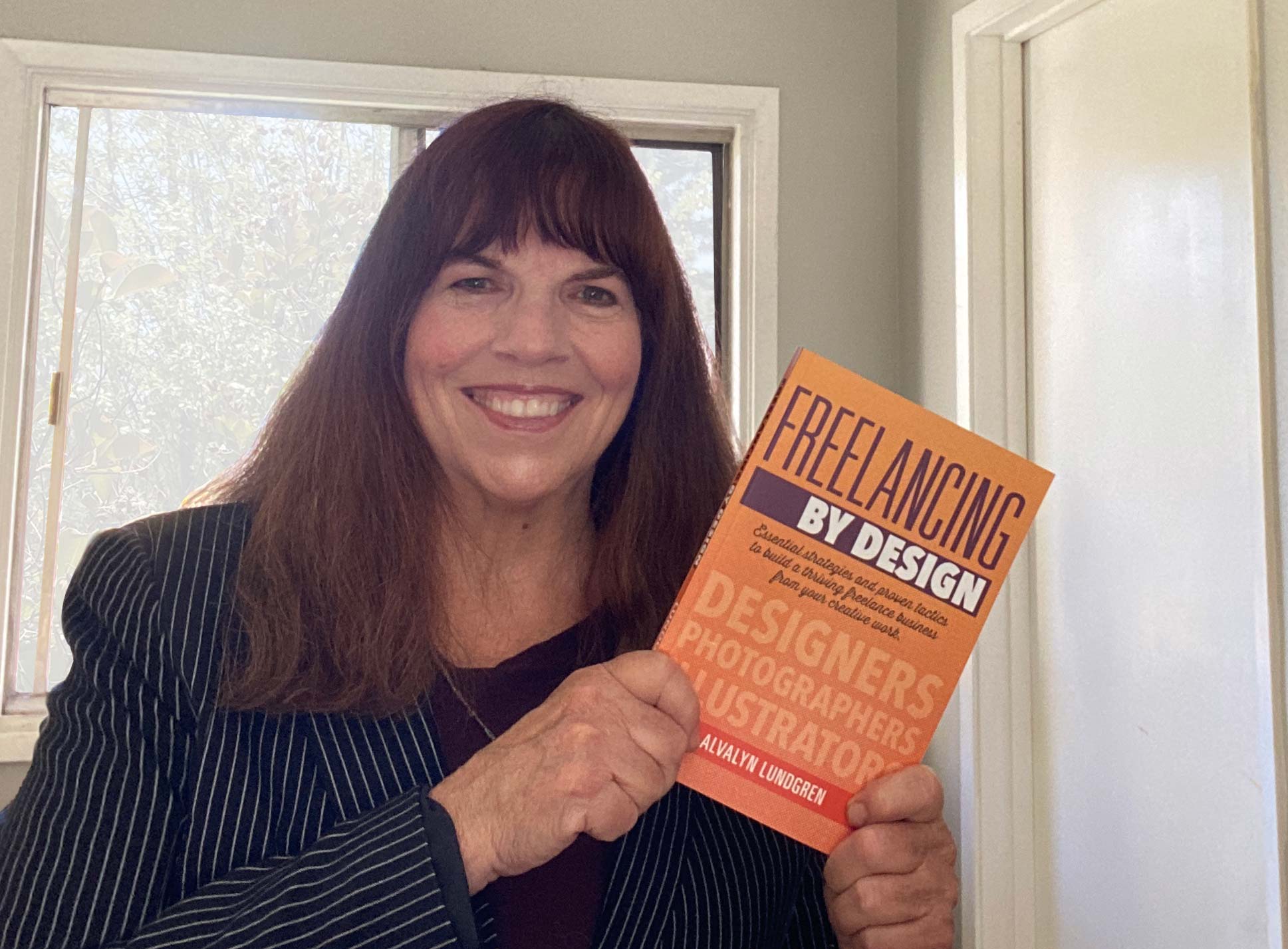 Woman with long dark hair smiling, holding a book titled 'Freelancing by Design' inside a room with a window.