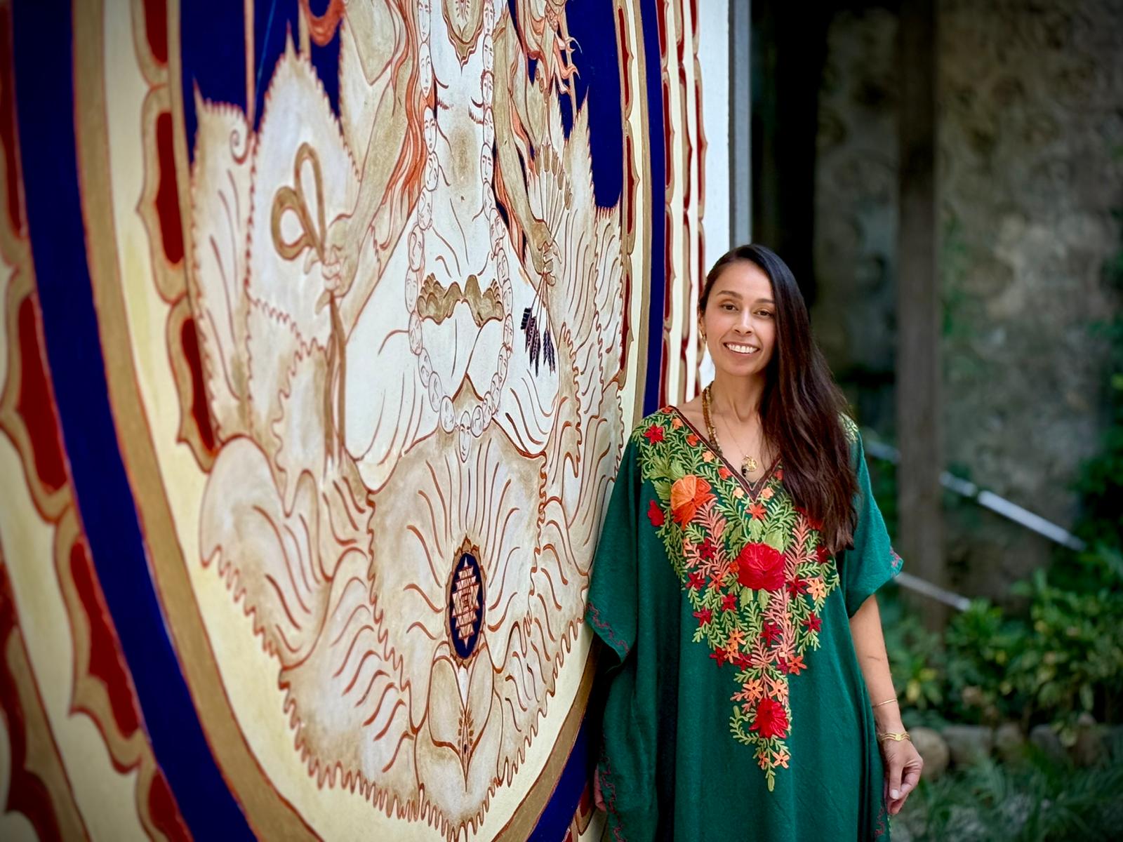Woman in green dress with red floral embroidery standing outdoors next to a large, colorful mural.