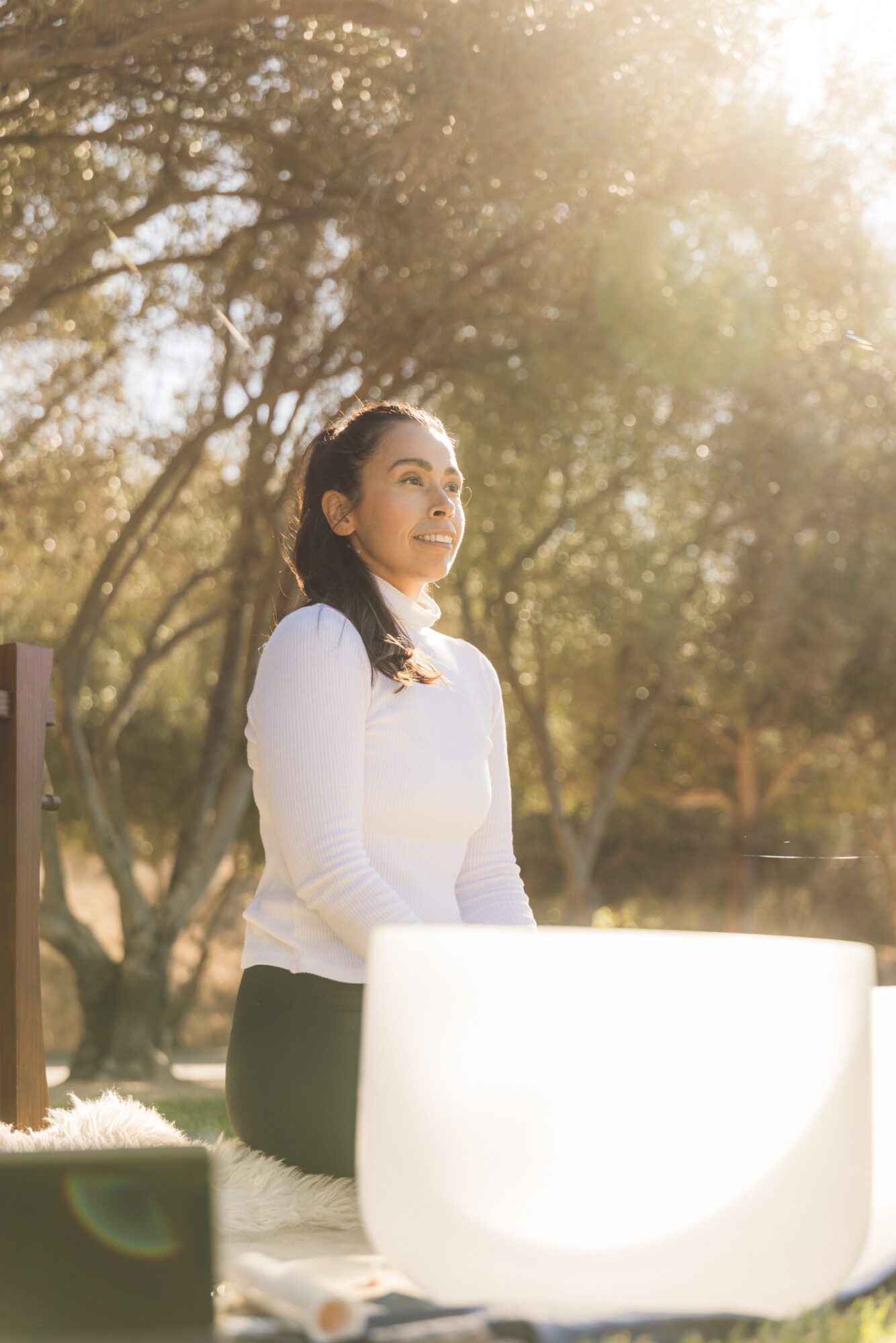Woman standing outdoors in sunlight, smiling, with trees in the background.