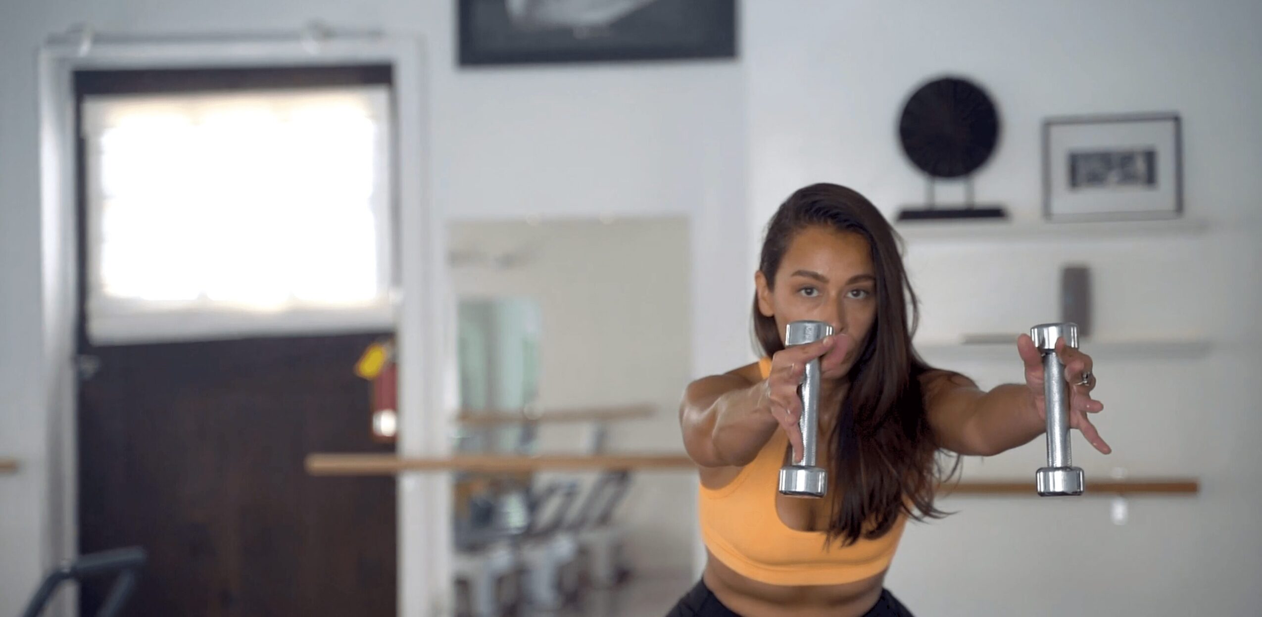 Woman in a gym holding dumbbells, reaching forward, with a window and shelves in the background.