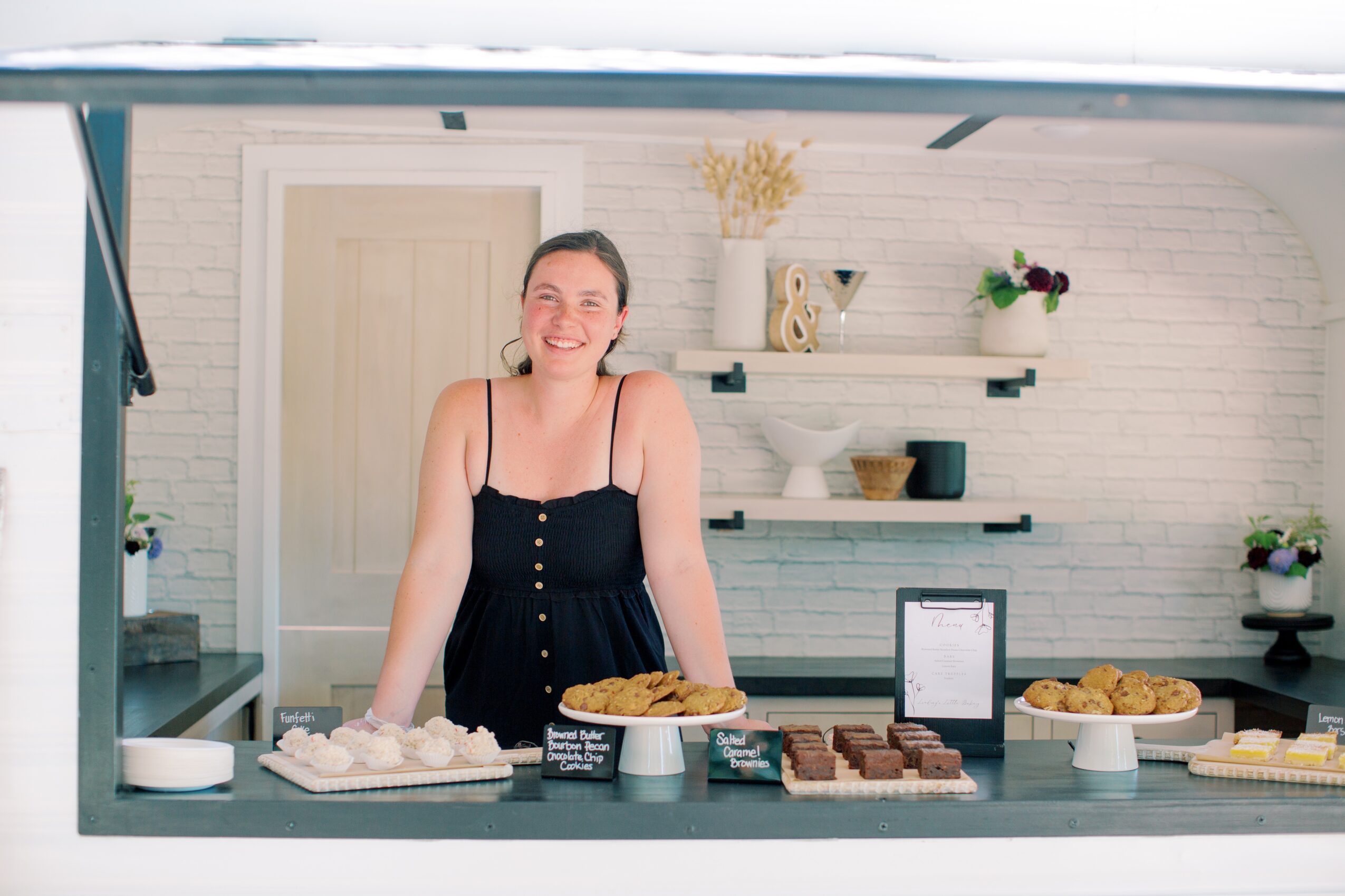 Smiling woman behind a table with baked goods, in a bright room with white brick wall and shelves.