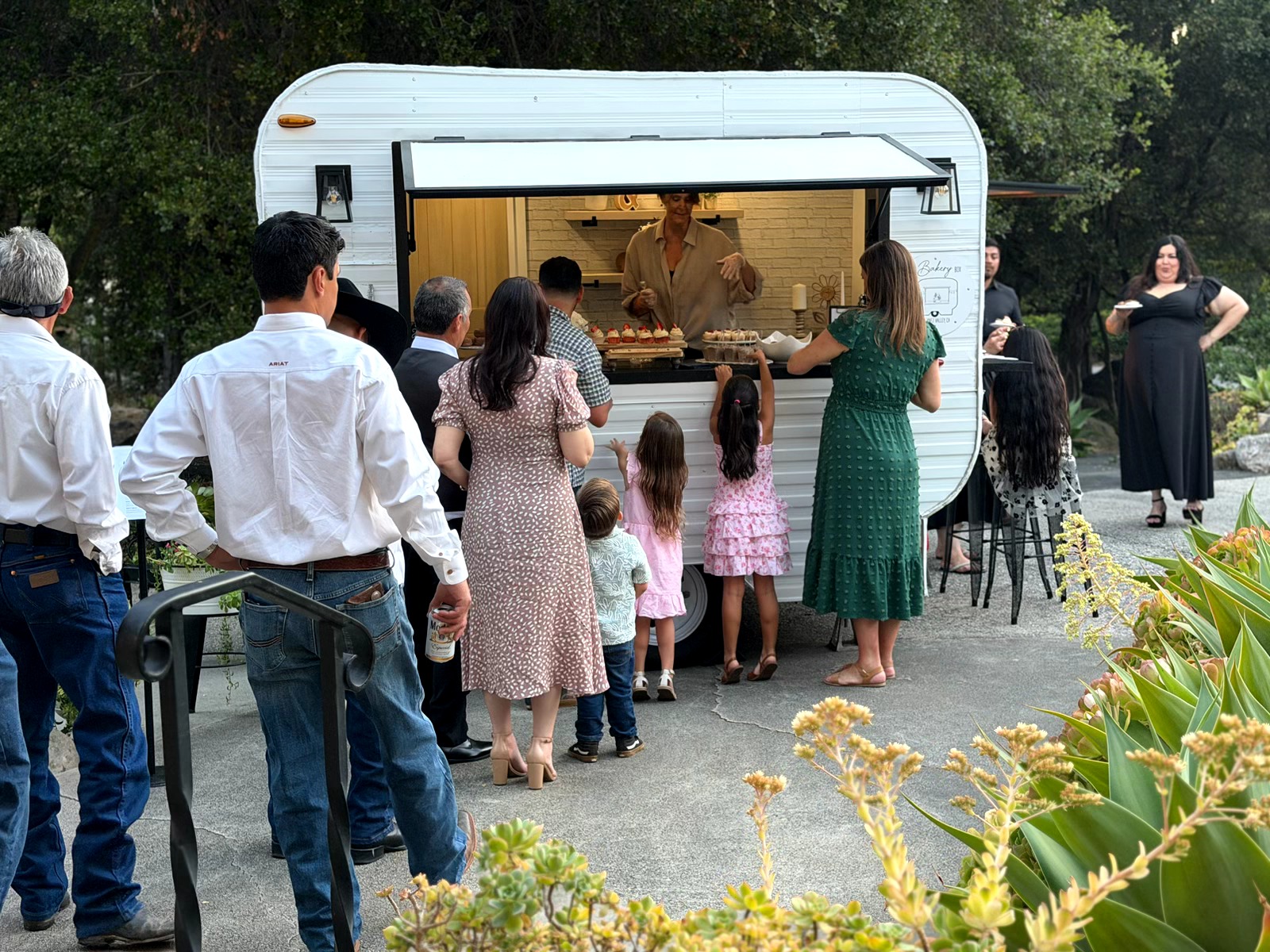 People gather around a food truck with a window serving food, outdoors with trees and plants nearby.