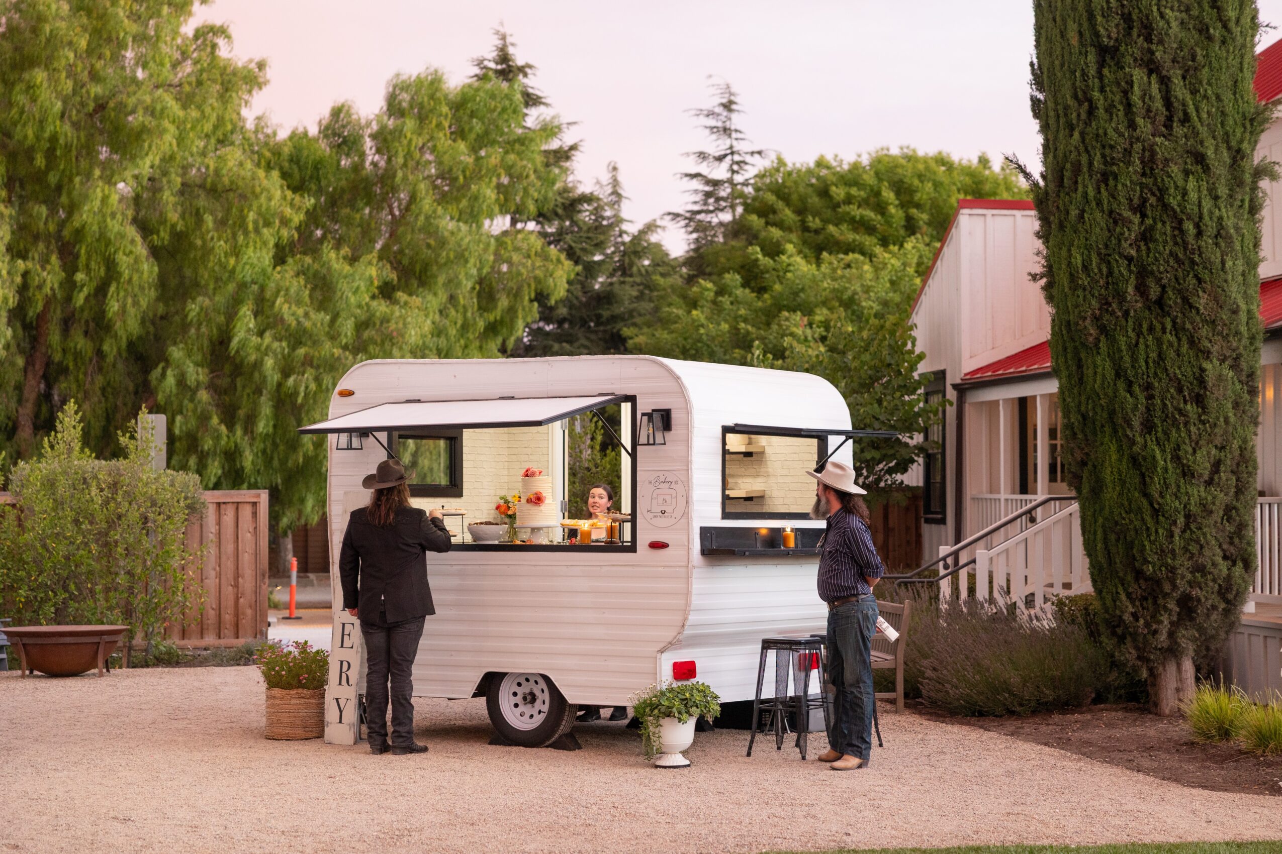 Two people stand outside a white food trailer with a window, surrounded by trees and a building, on a gravel surface.