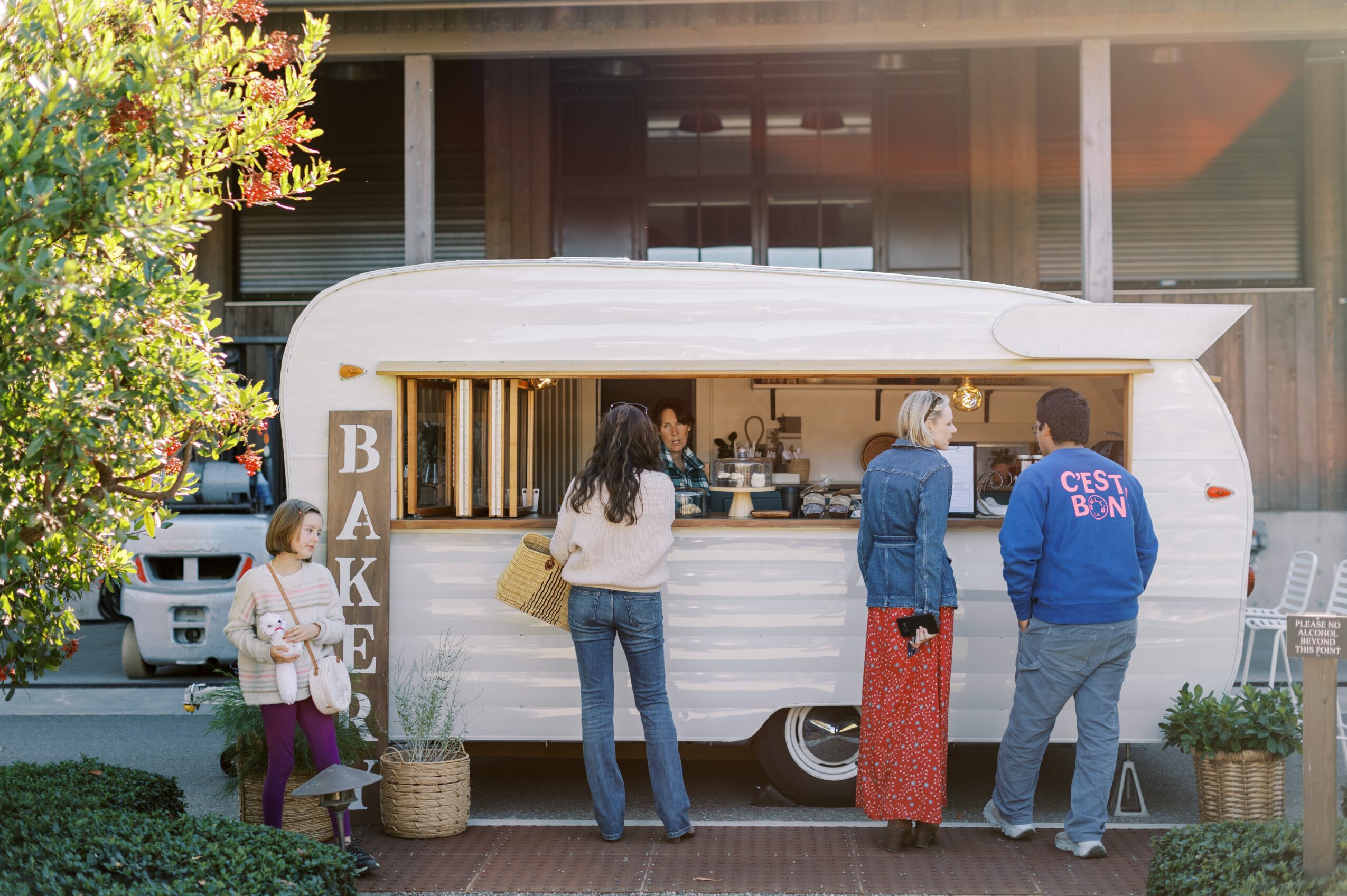 People order food at a white food truck with a menu, outside a building with large windows, trees, and parked cars.