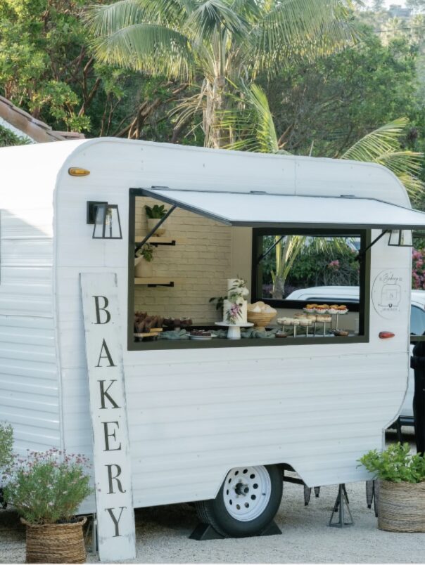 A white bakery trailer with a large window displaying baked goods, outdoors with green trees in background.