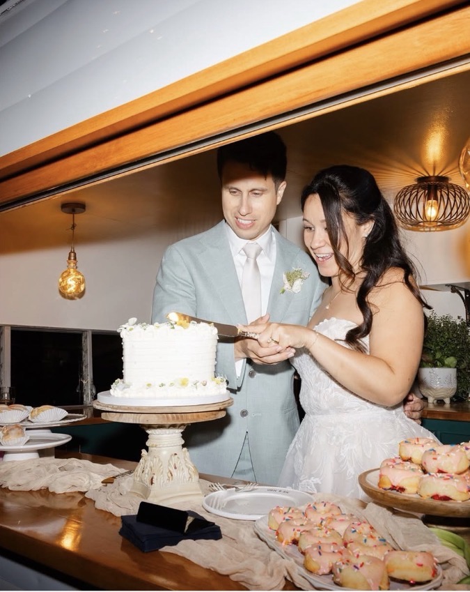 A couple in wedding attire cutting a cake at a celebration, with desserts on the table and warm lighting in the background.