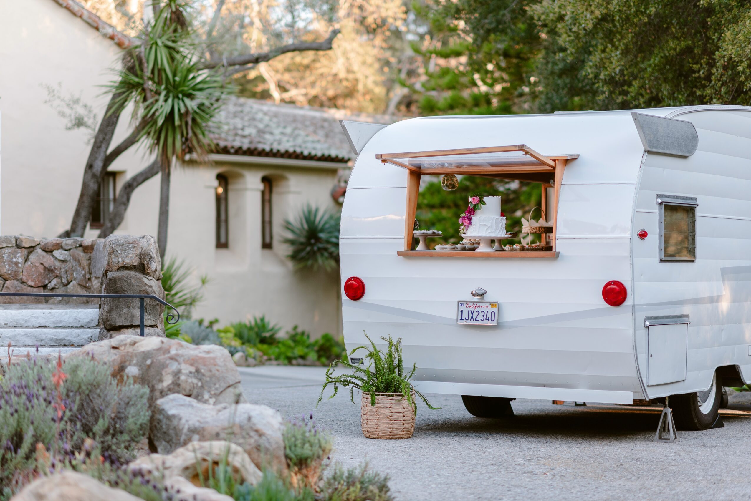 White camper trailer parked outdoors with plants and rocks nearby, a window with a wooden frame, and a license plate reading 'LAAZ'.