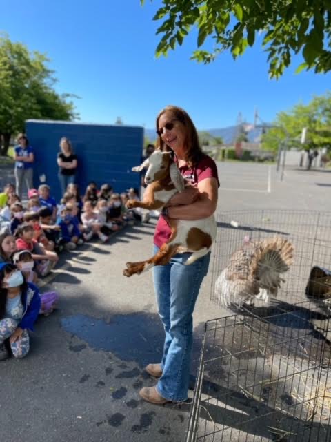 Woman holding a dog in front of children sitting outdoors, with a caged dog nearby and trees in background.