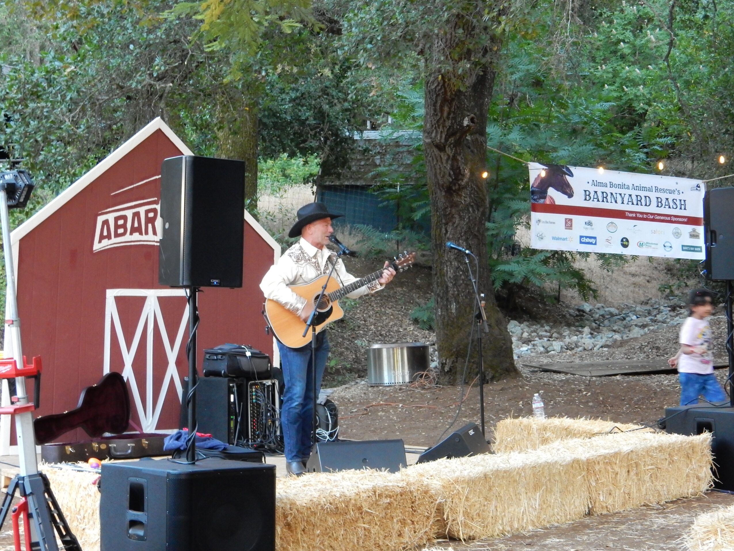 Child playing guitar on stage with hay bales, trees, and banners in background, outdoor event setting.