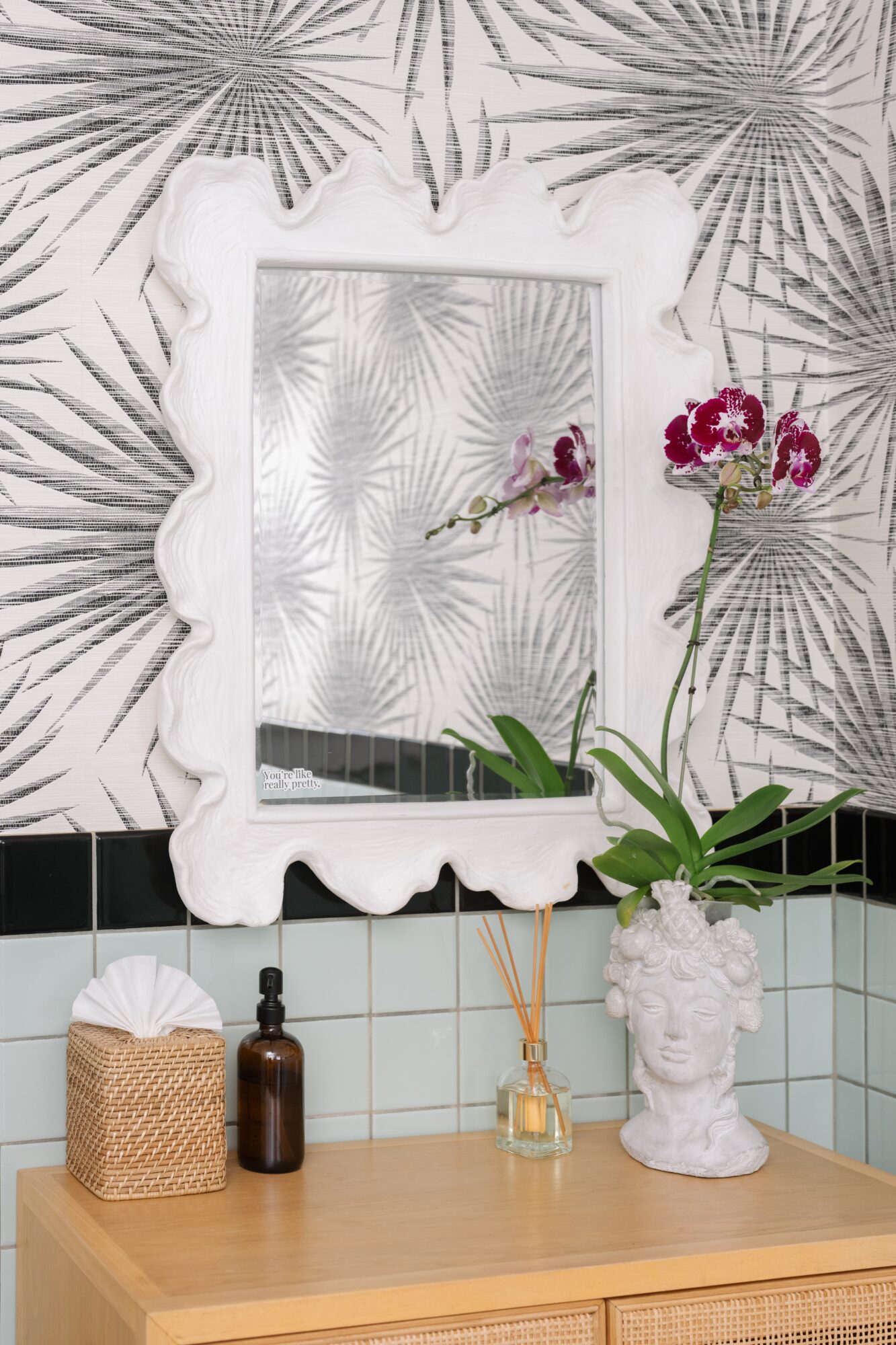 Decorative mirror with a white ornate frame on a wall with palm leaf wallpaper, above a wooden surface with plants and decor.