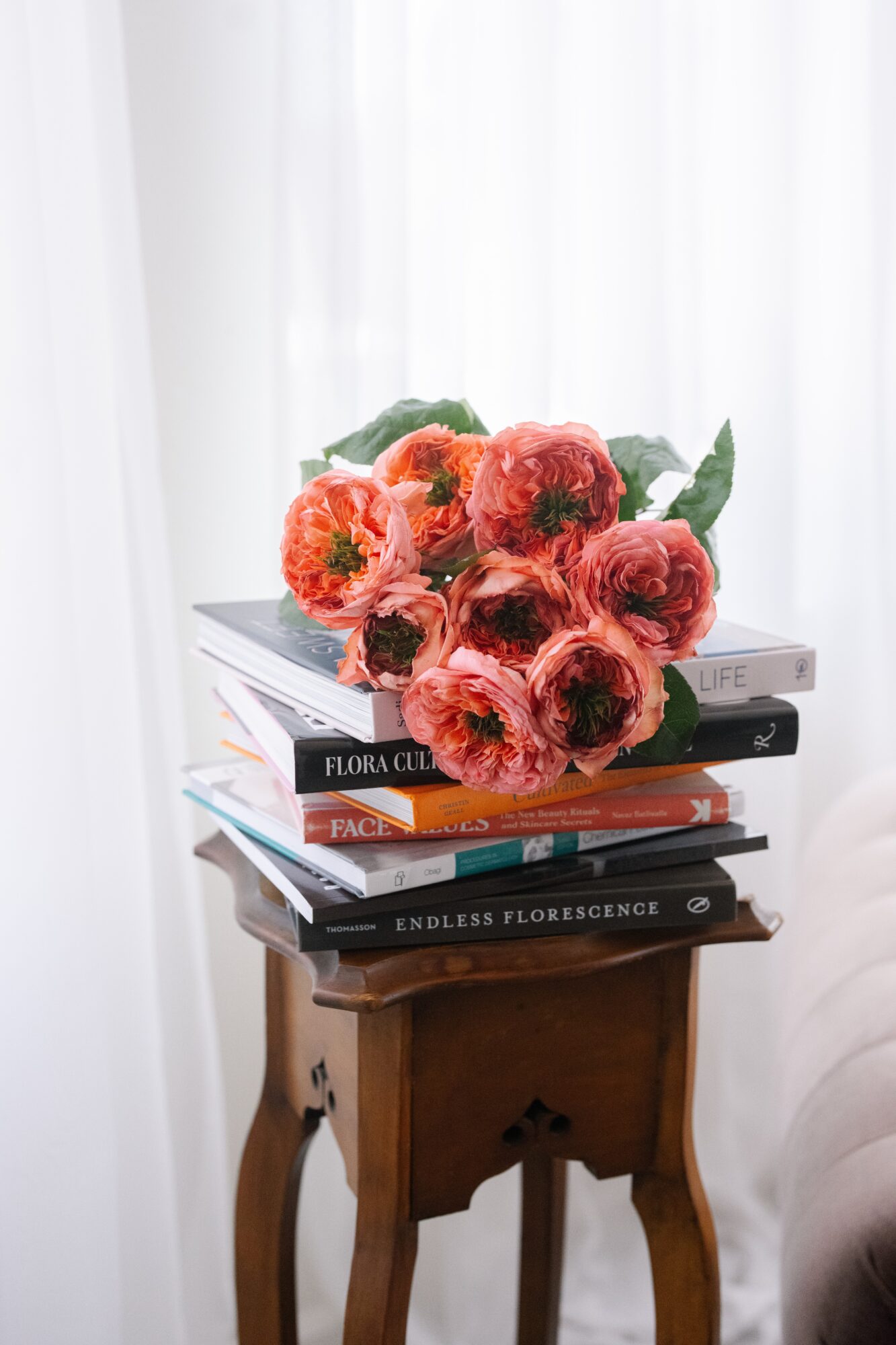 Pink and orange peony flowers on a stack of books on a small wooden table, with a white curtain background.