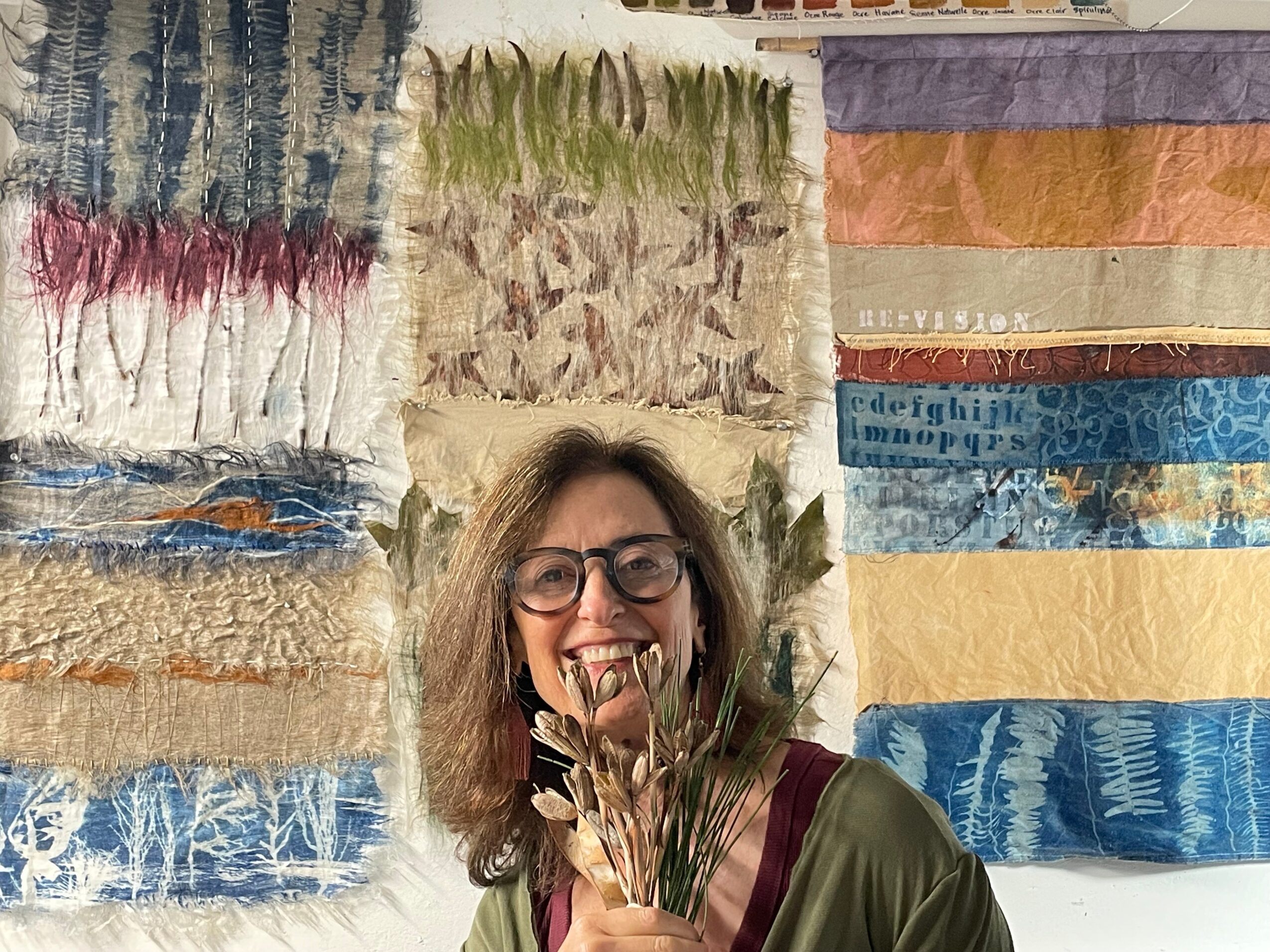 Woman with glasses smiling, holding plants, with colorful fabric or textile background.