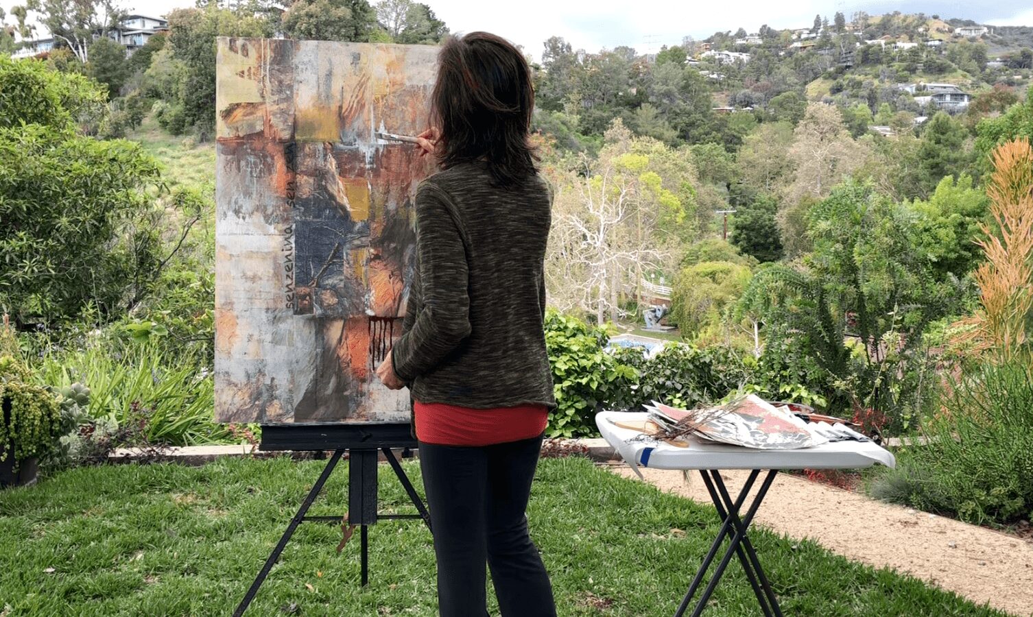Person standing outdoors in front of an abstract painting on an easel, with trees and hills in the background.