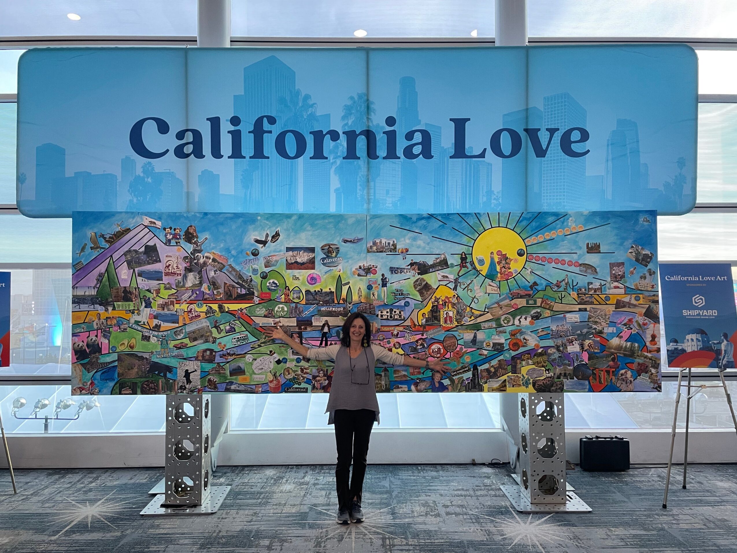 Person standing in front of a large sign that reads 'California Love' with a colorful mural below, city skyline in background.