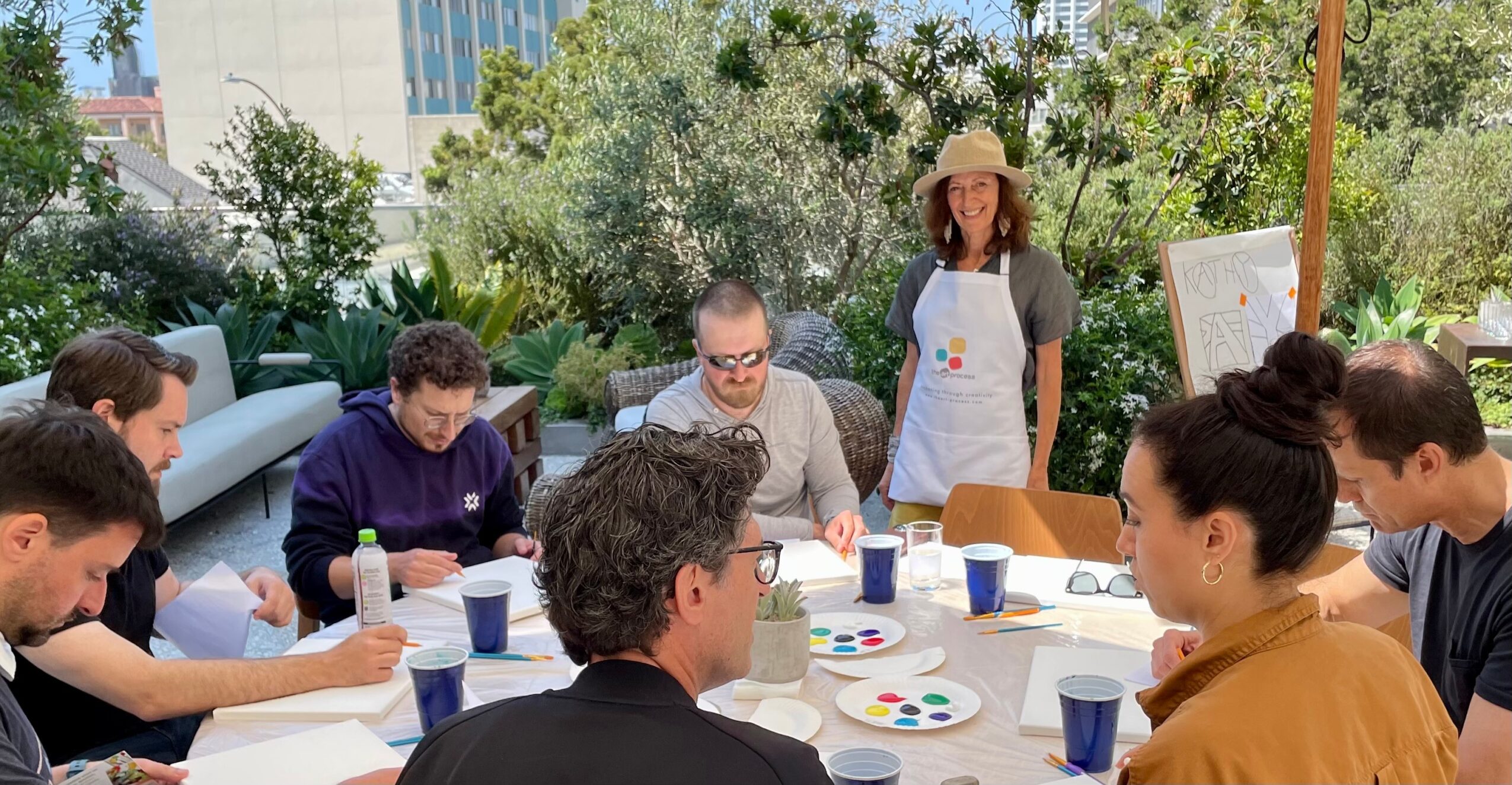 Group of people sitting around a table outdoors, painting and drawing, with a woman standing nearby, trees and buildings in background.