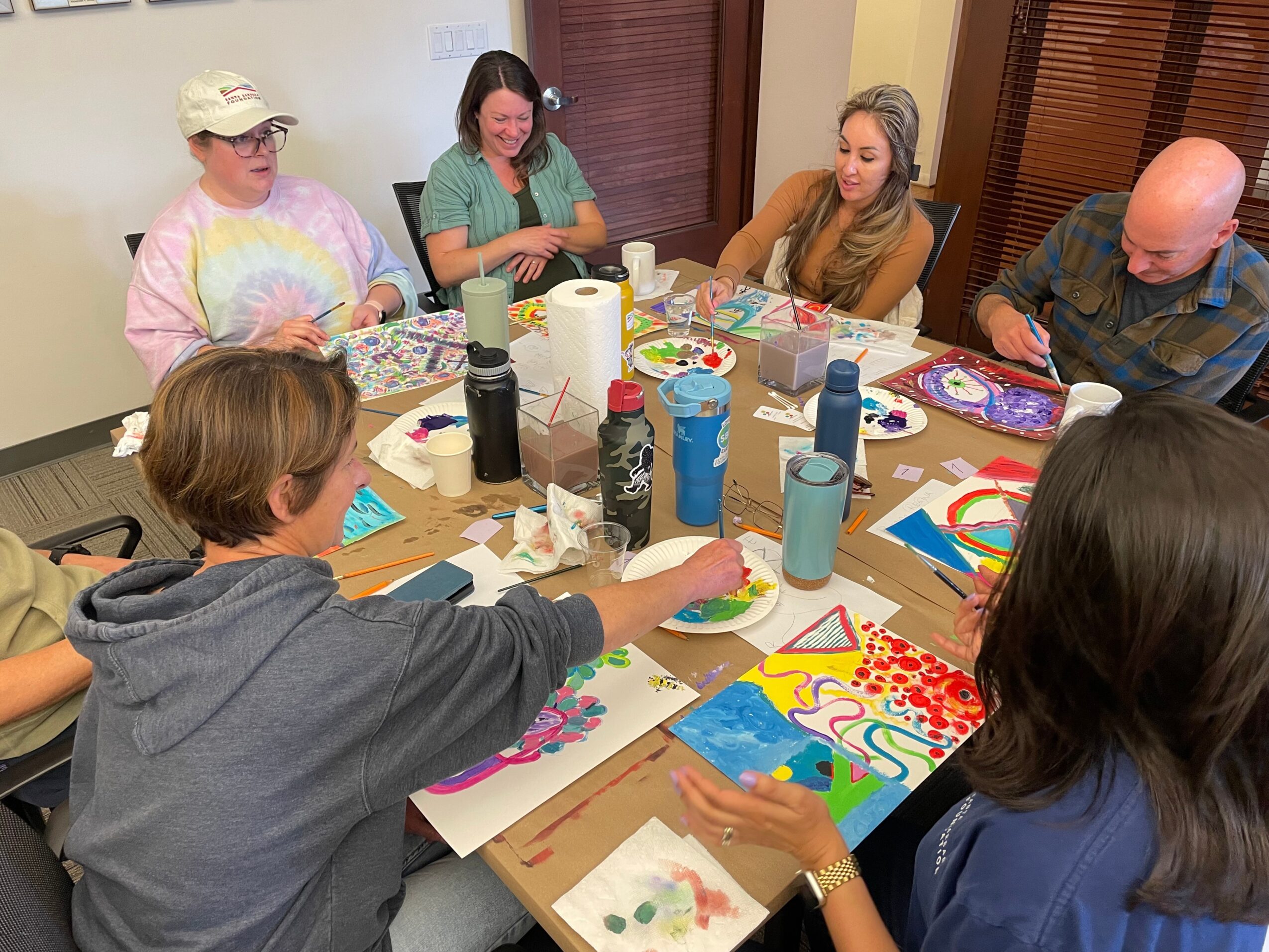 Group of six people sitting around a table with art supplies, creating colorful artwork in a room.