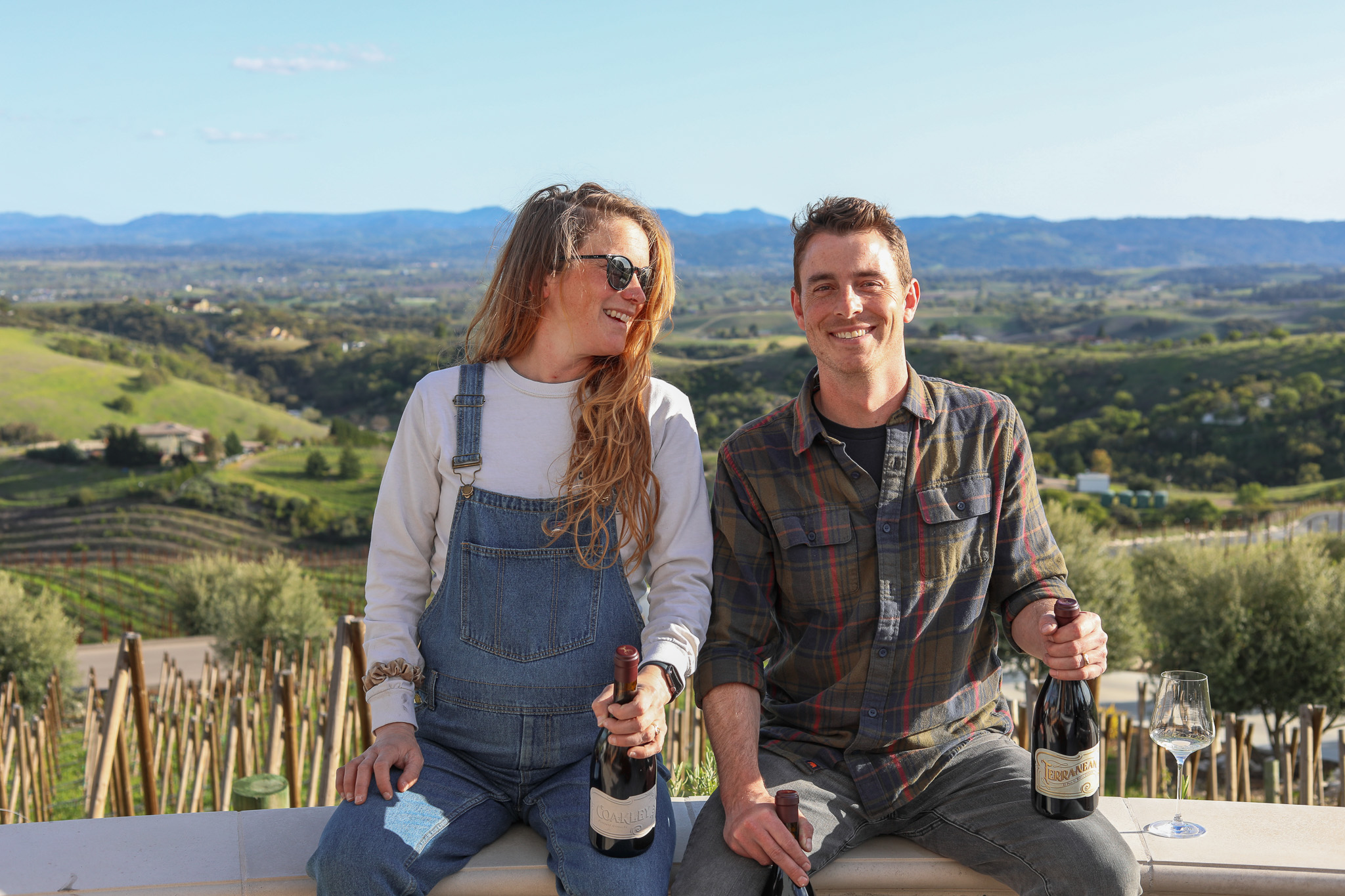 A young man and woman stand outdoors with a scenic landscape in the background, holding bottles of wine, smiling.