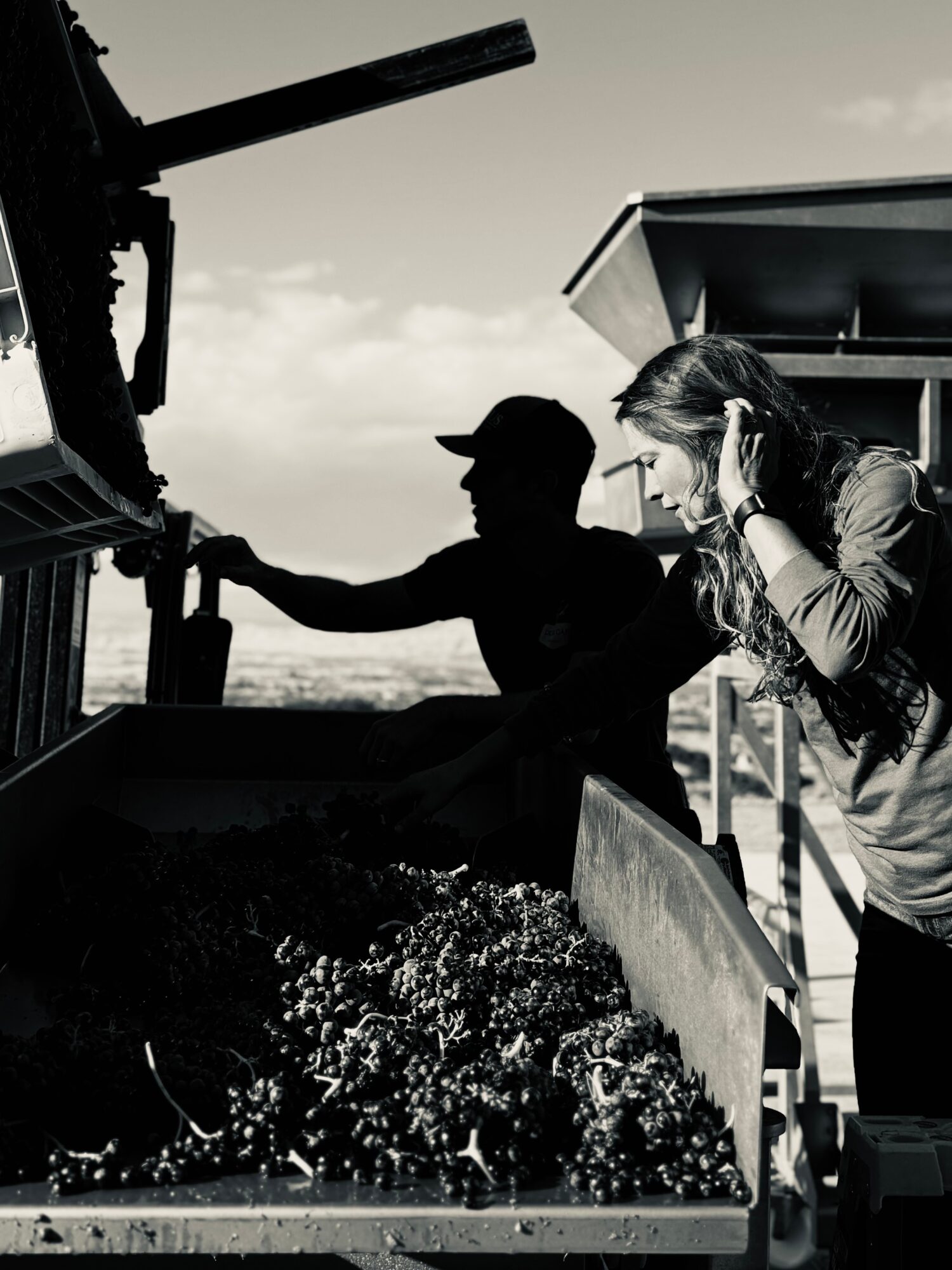 Two people working outdoors near a large container, with one person wearing a cap and the other with long hair, in black and white.