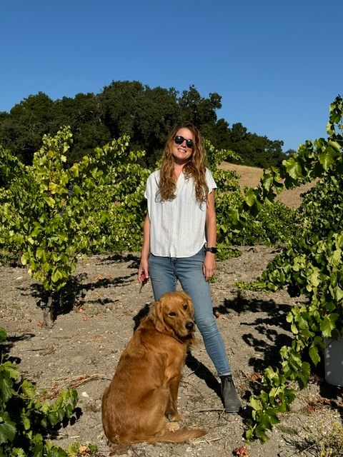 Woman with sunglasses standing outdoors next to a sitting golden retriever, surrounded by green plants and trees under a blue sky.