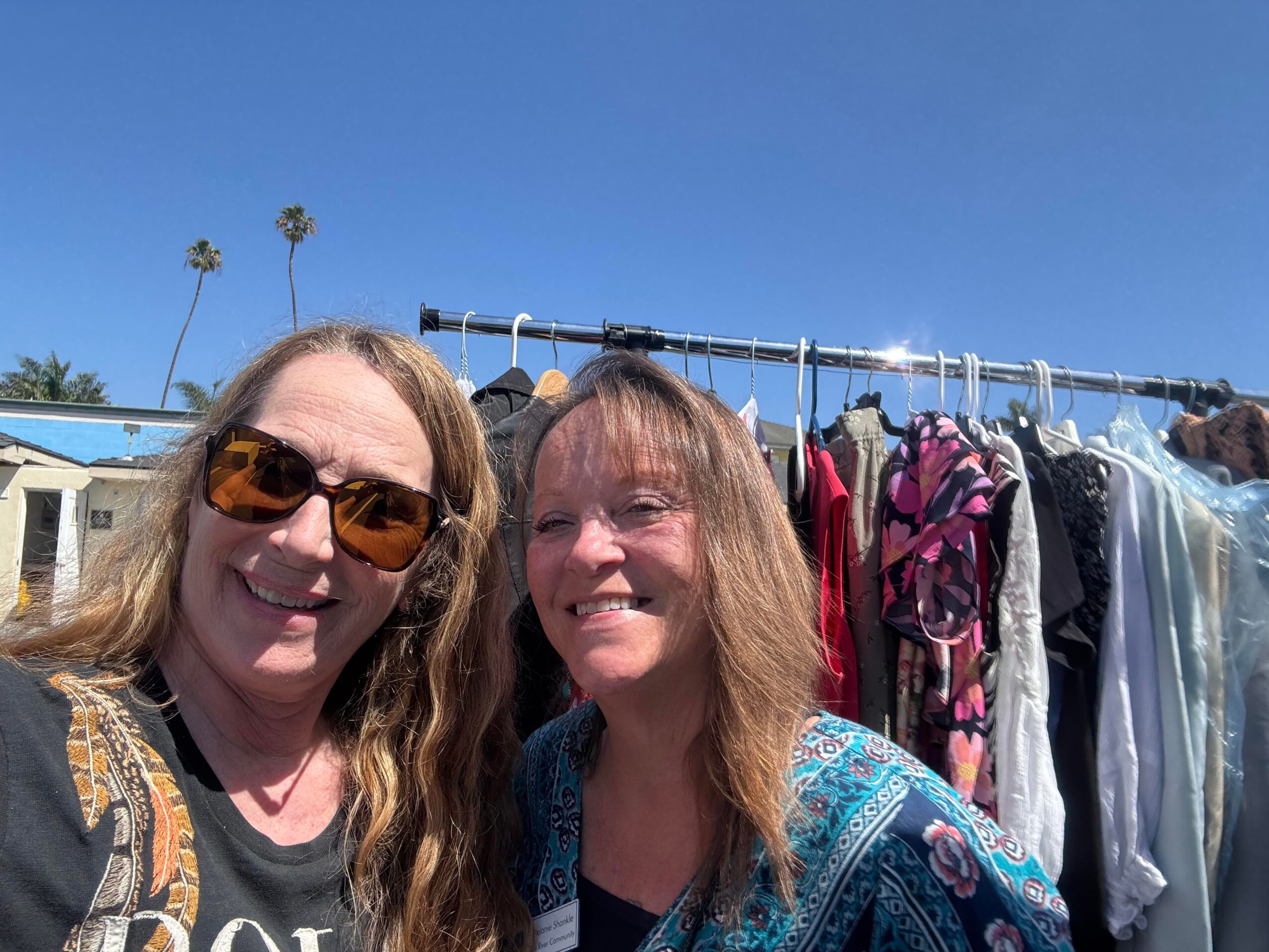Two women smiling outdoors with a clothing rack and blue sky in background.