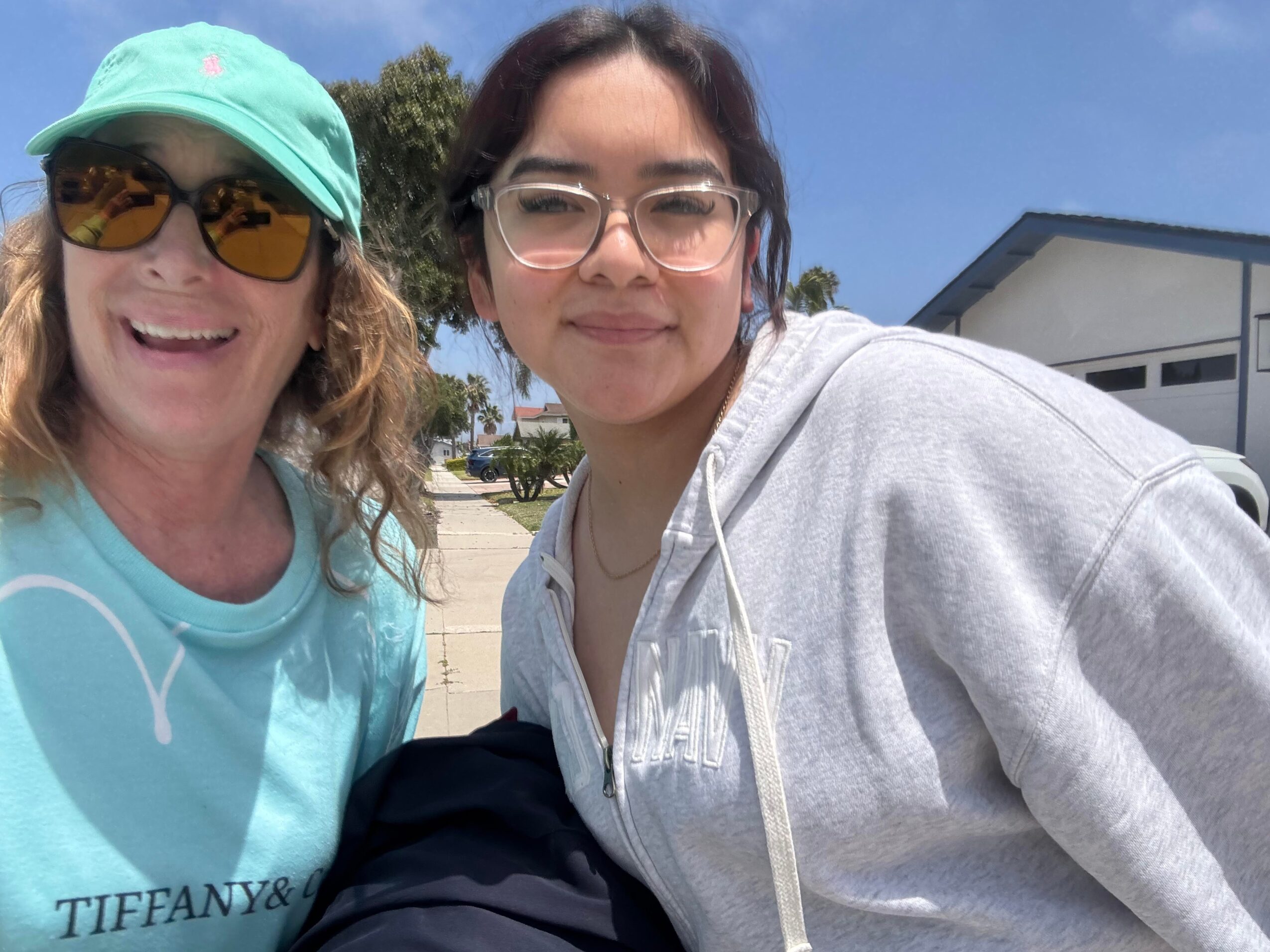 Two women smiling outdoors, one wearing a turquoise cap and sunglasses, the other with glasses and a gray hoodie.