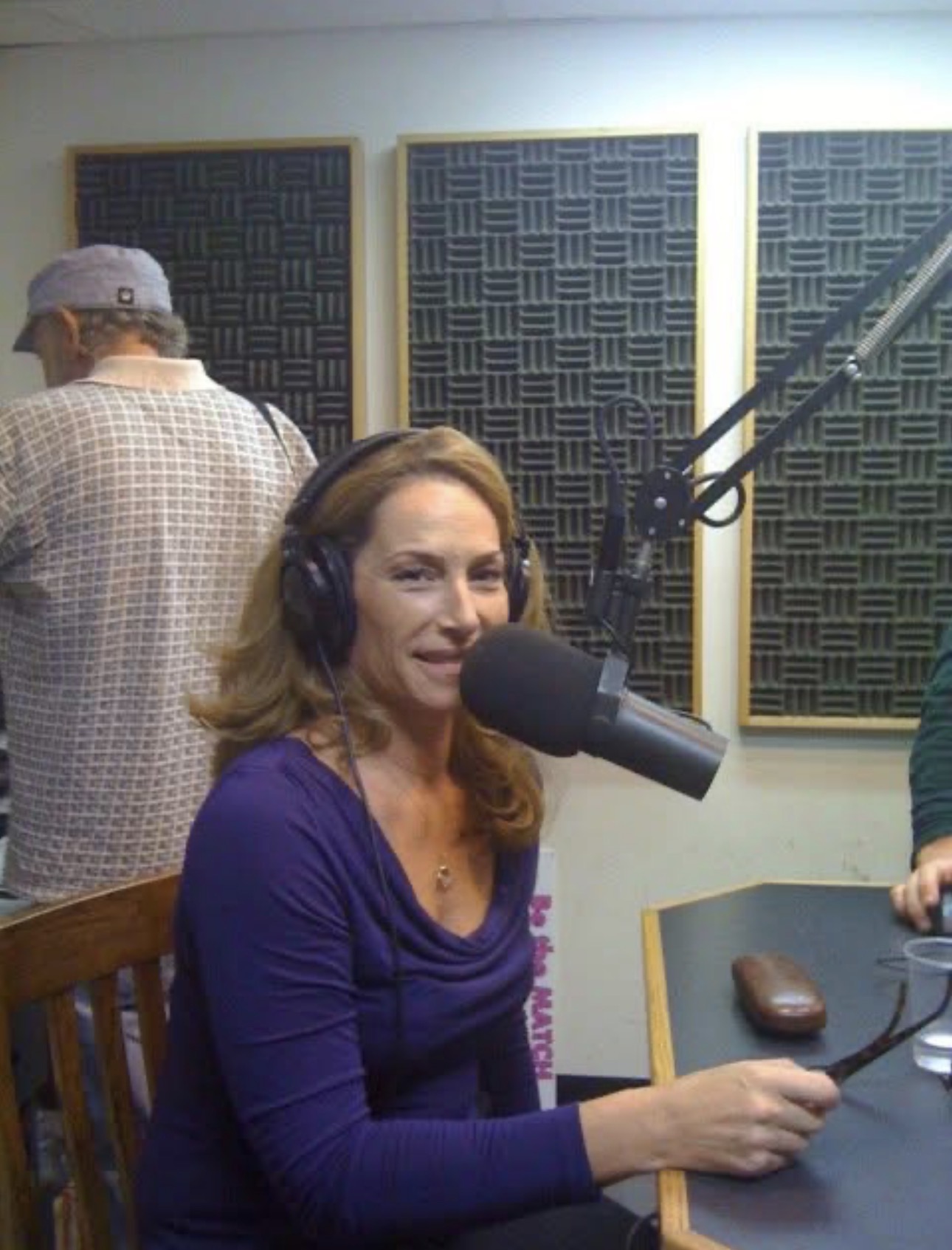 Woman with headphones speaking into microphone in recording studio, with person in background wearing a hat, acoustic panels on wall.