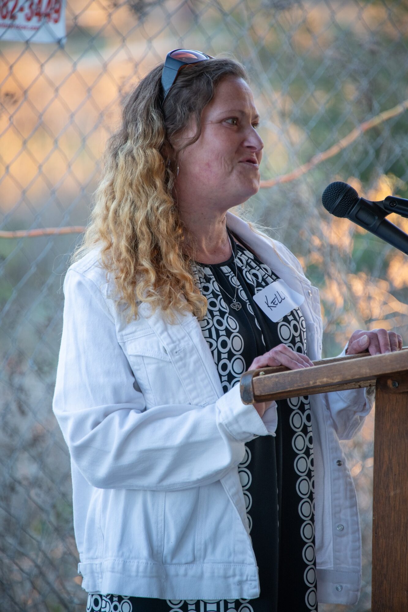Woman with curly hair speaking at a podium outdoors, wearing a white jacket and patterned top, with a microphone nearby.