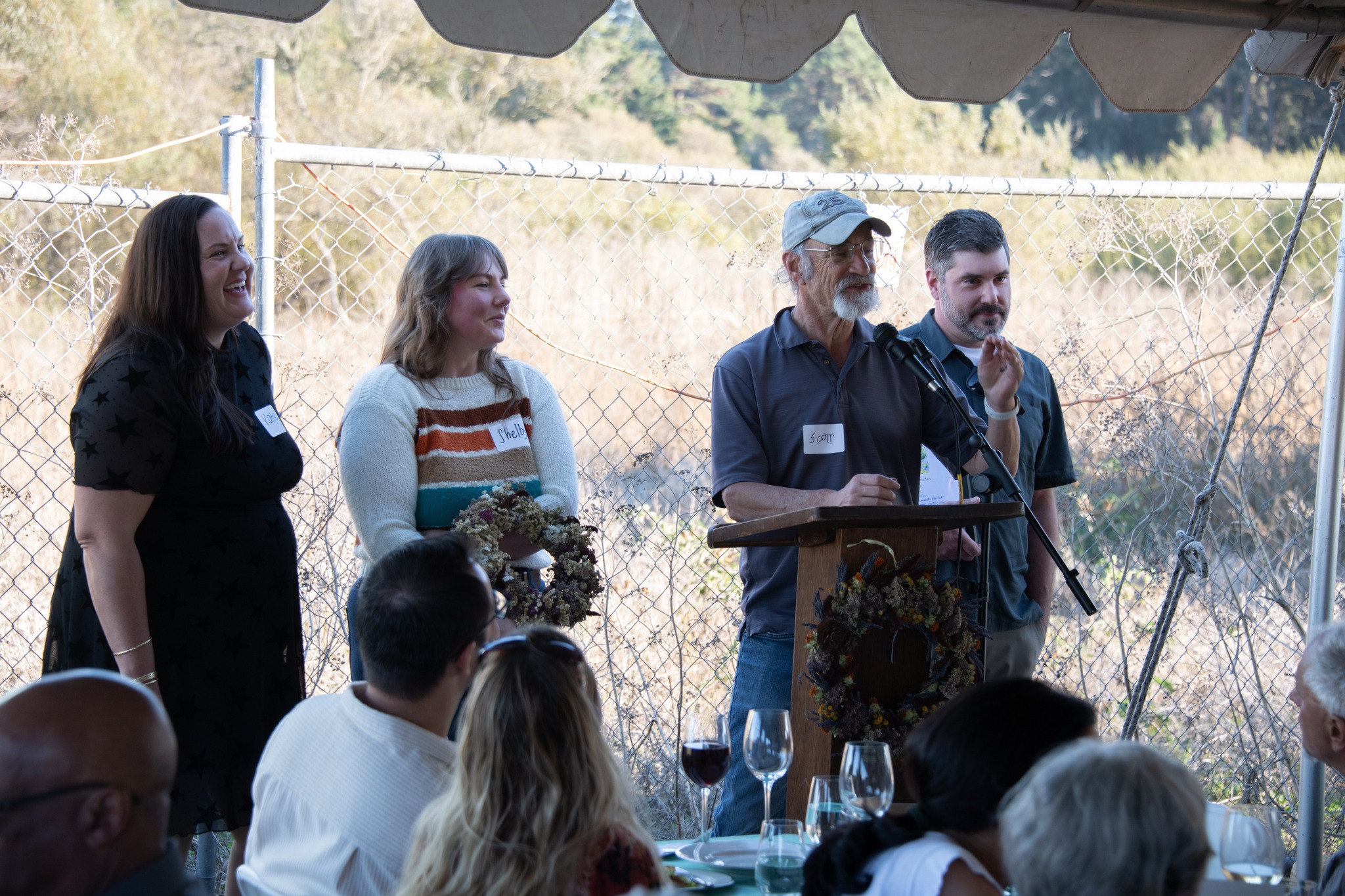 Four people standing outdoors under a canopy, speaking at a podium with a microphone, with an audience in front, fence in background.