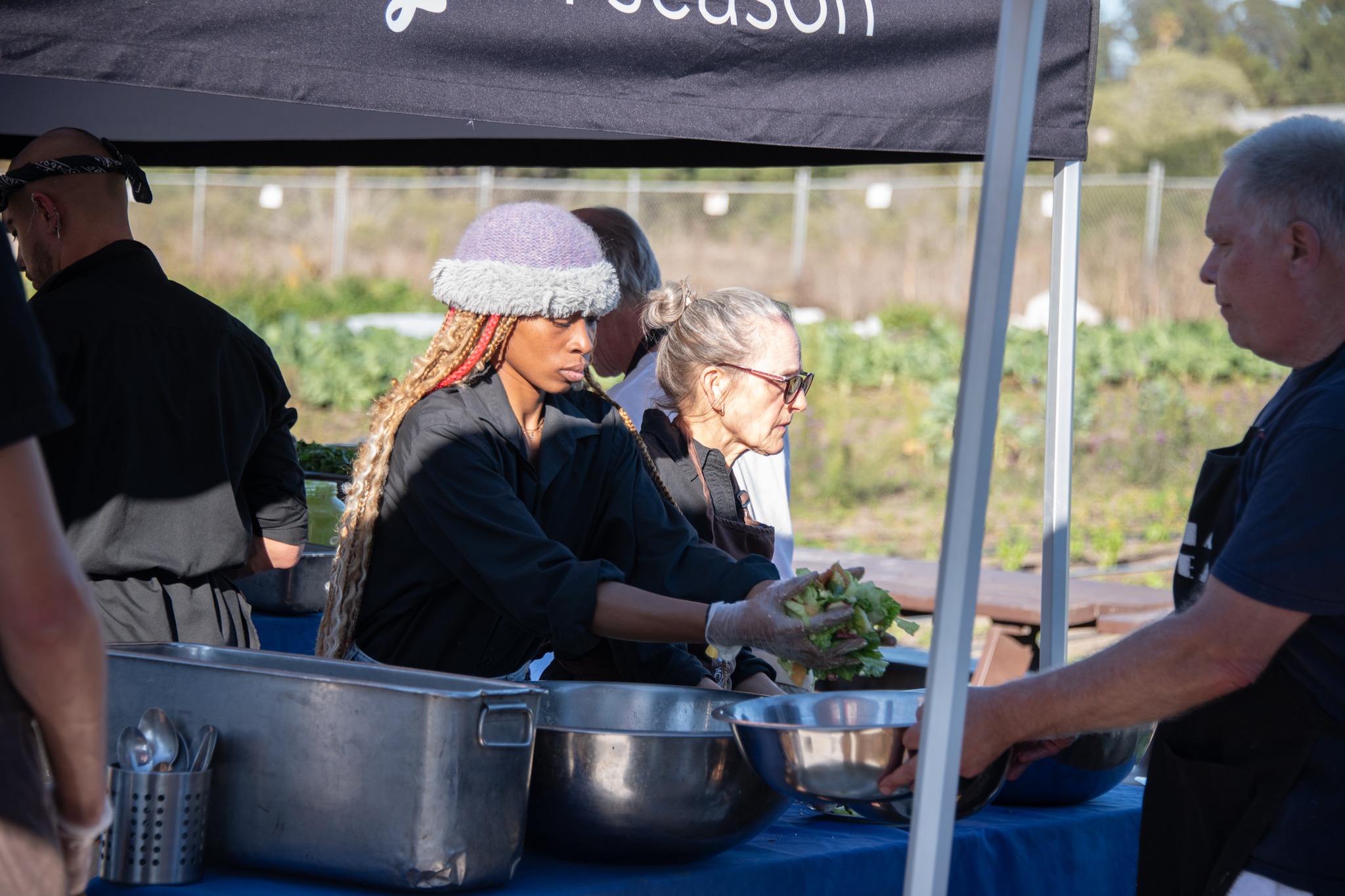 Two women and a man at an outdoor food stand, with a tent overhead, preparing food with bowls and utensils.