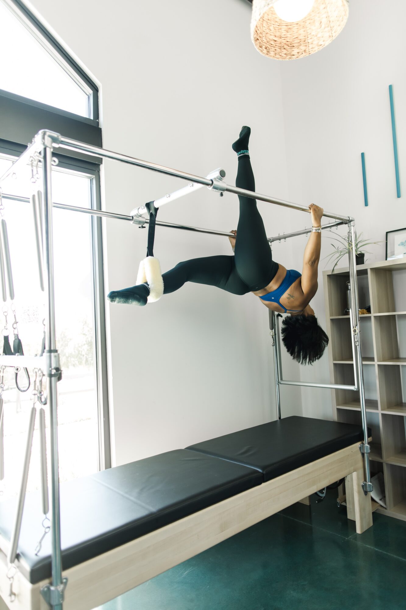 Person performing aerial move on horizontal bar in a room with white walls and a window.