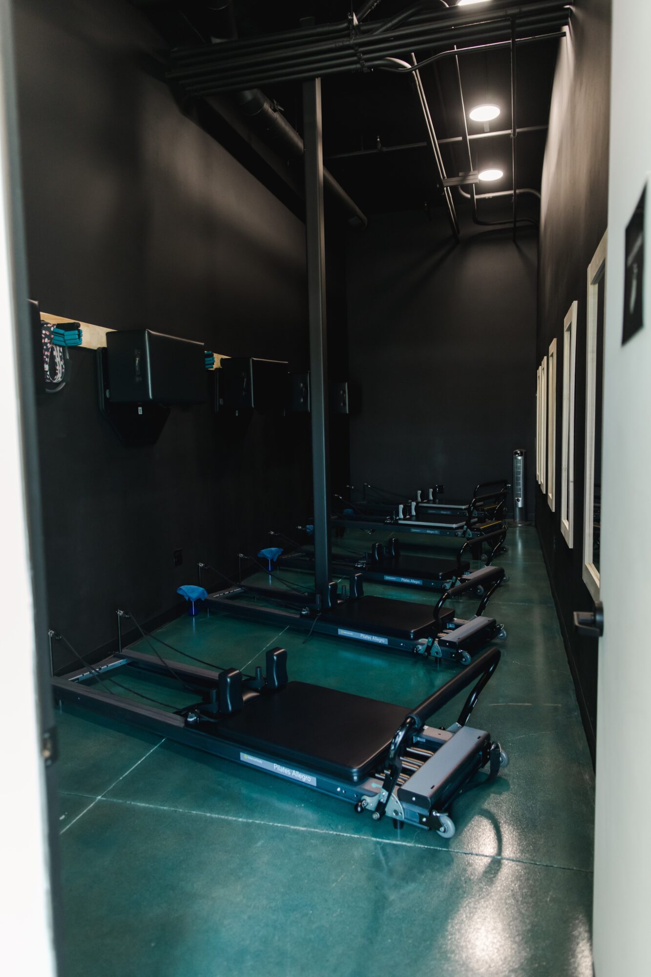 Row of exercise machines in a dark gym with black walls and ceiling, illuminated by ceiling lights.