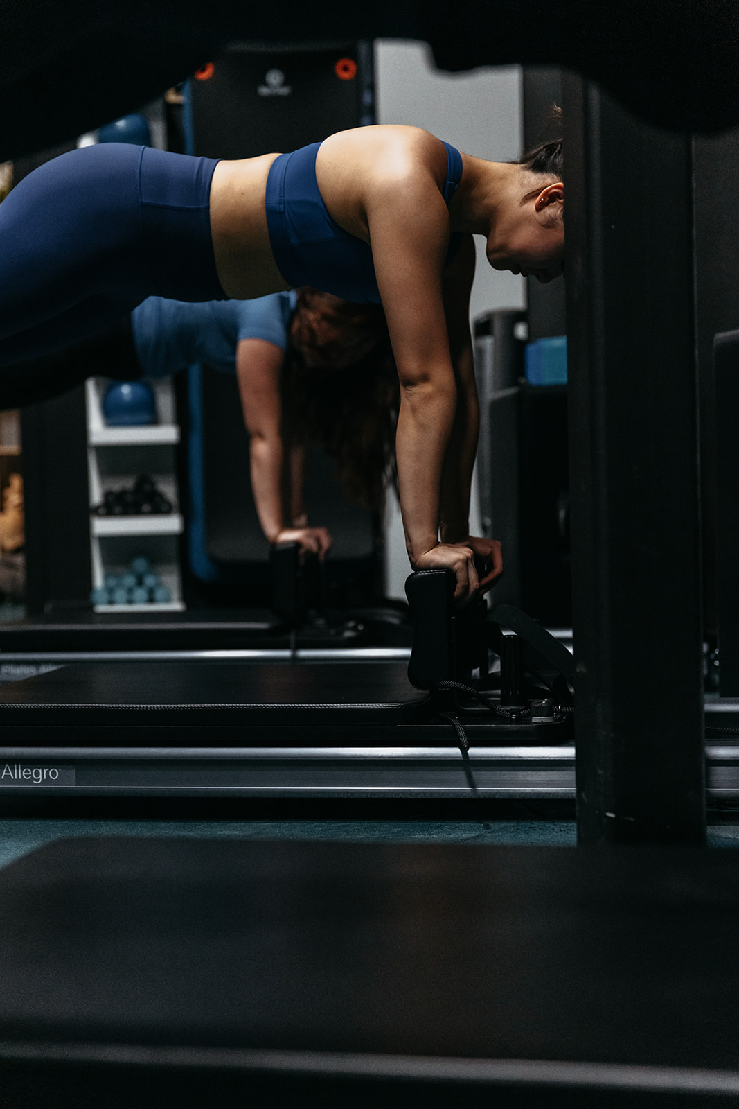 Woman in workout clothes performing a plank with hands on dumbbells in a gym, with a person in the background. Word count: 20