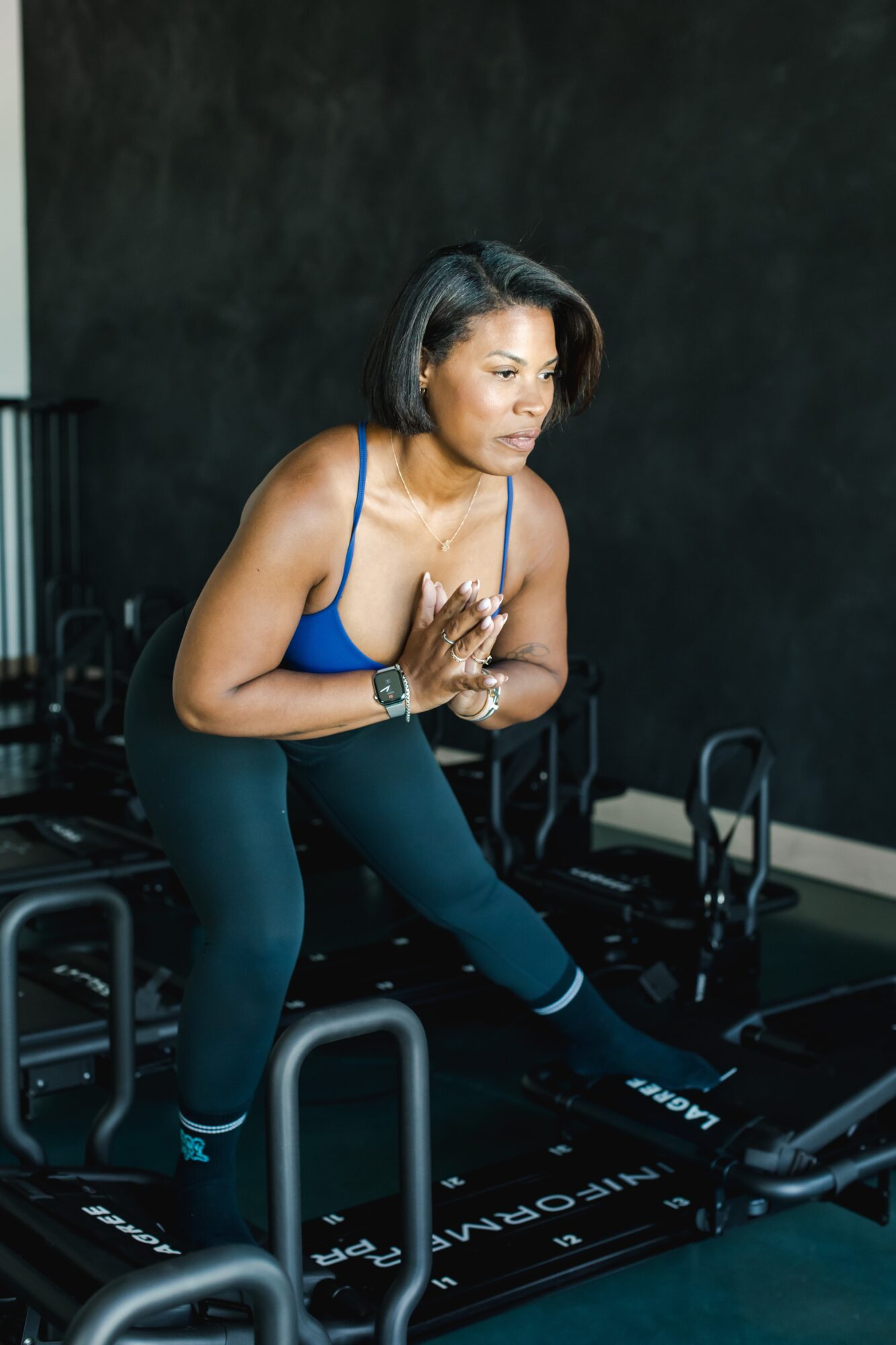 Woman in workout clothes on a treadmill, hands pressed together in a prayer position, in a gym setting.
