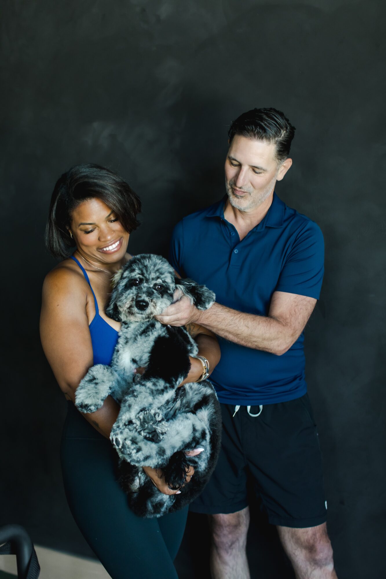 A woman and a man hold a large, fluffy puppy against a dark background.