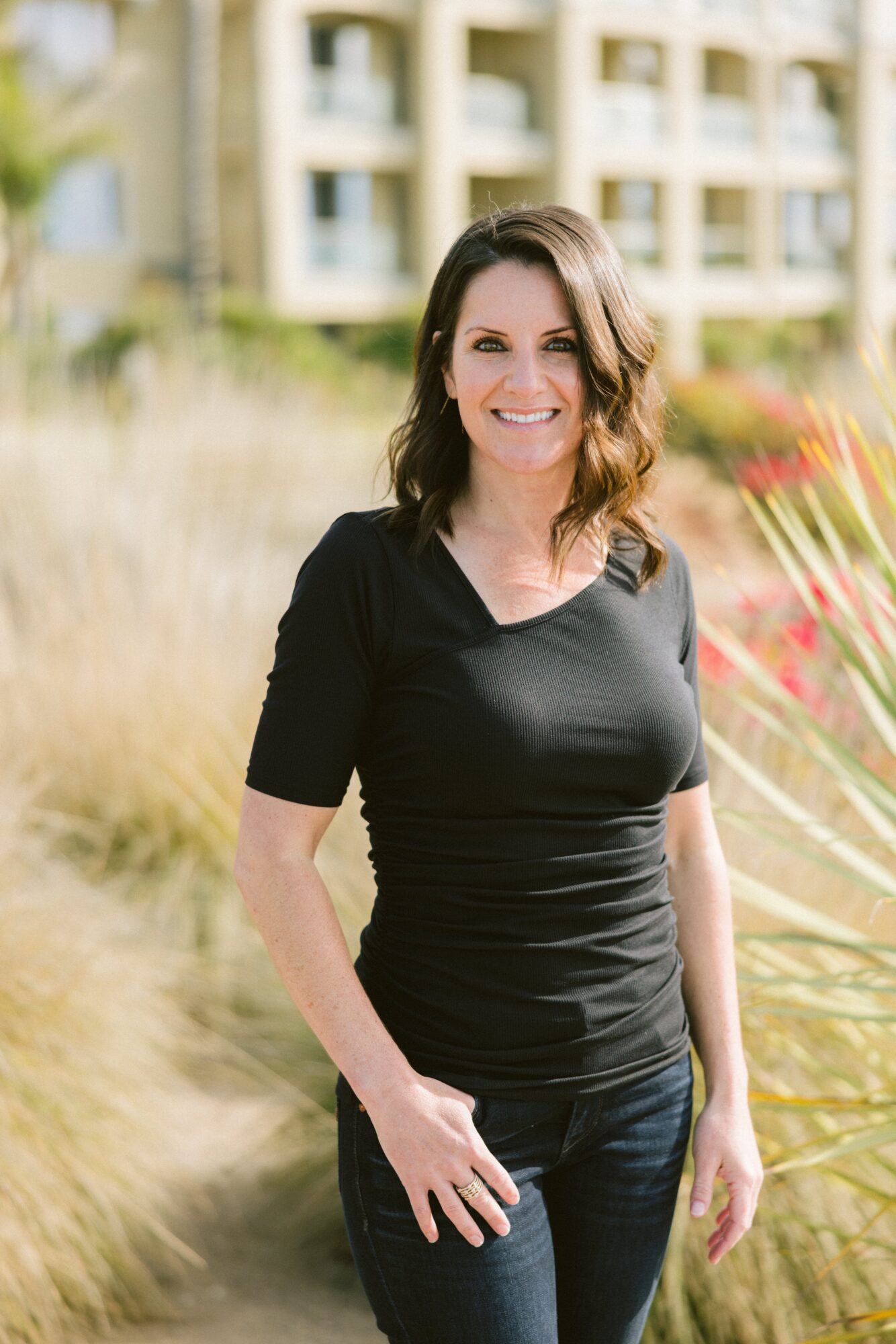 Woman with shoulder-length hair smiling outdoors, wearing a black top and jeans, with buildings and plants in background.