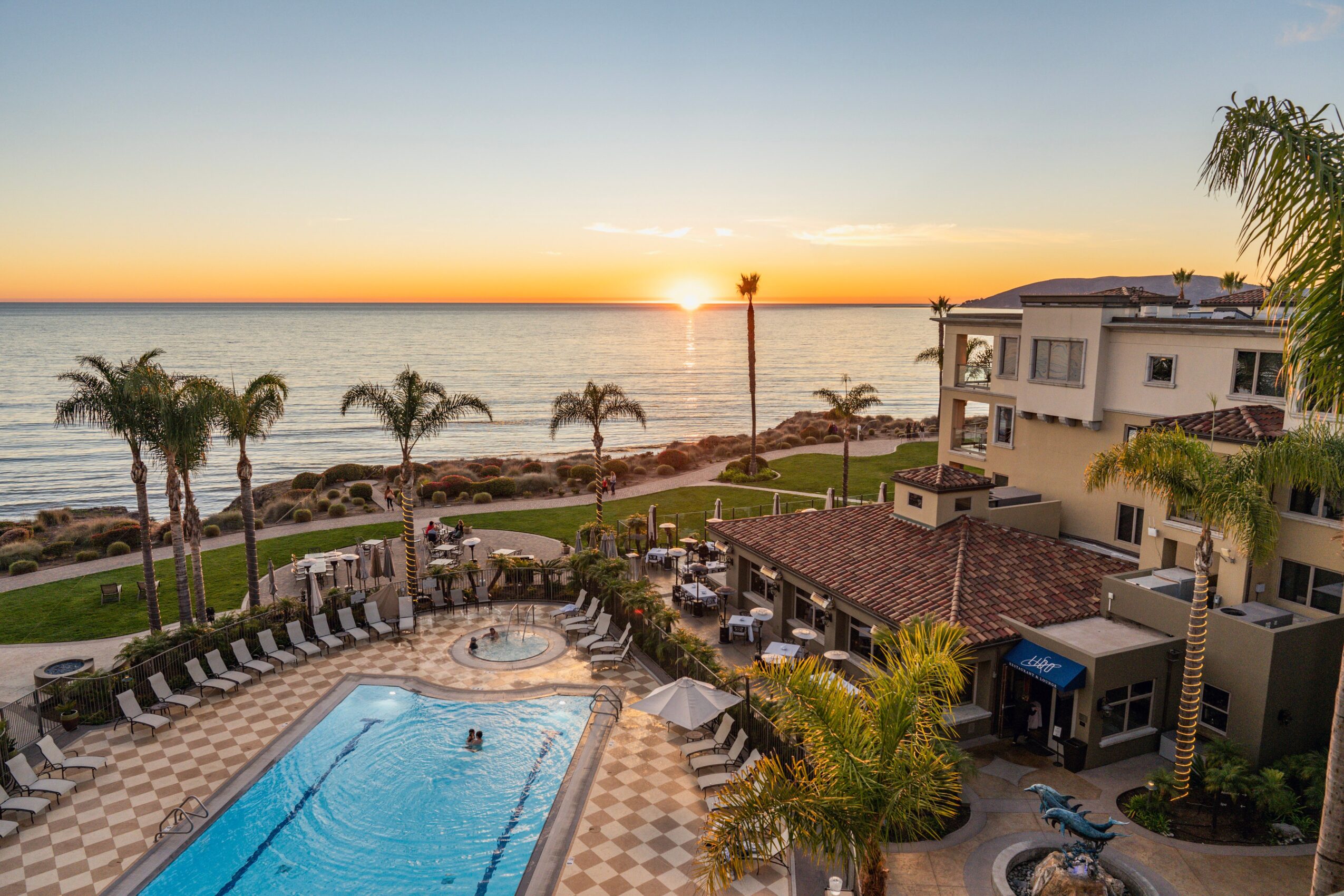 Pool area with lounge chairs, palm trees, and a view of the ocean at sunset, with buildings nearby and a clear sky.