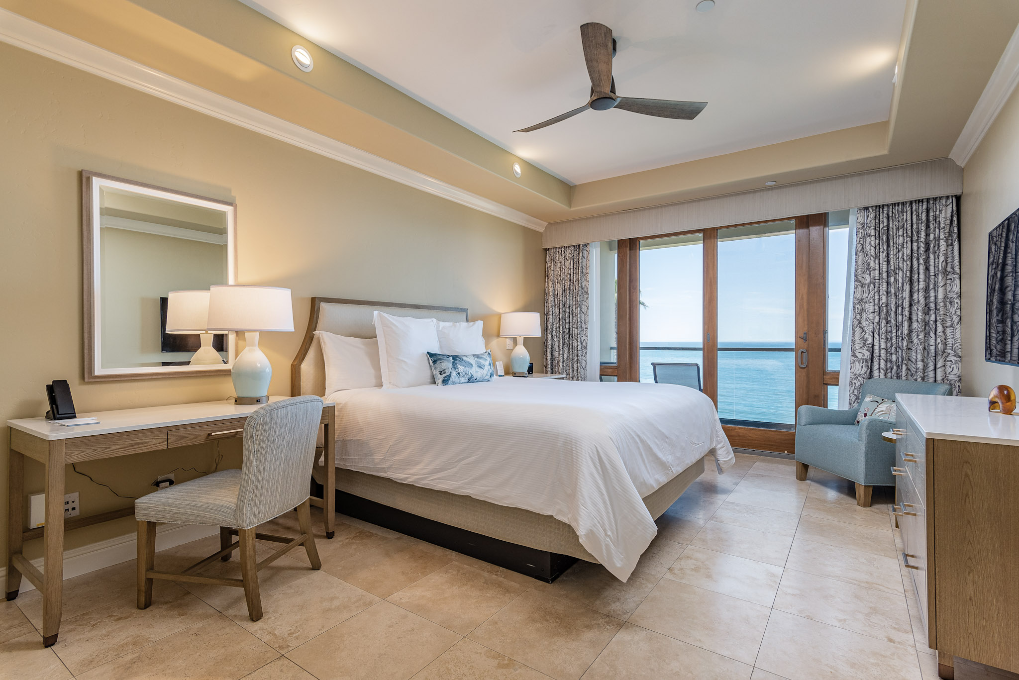 Hotel room with a large bed, desk, mirror, armchair, and balcony with ocean view, bright and airy space.