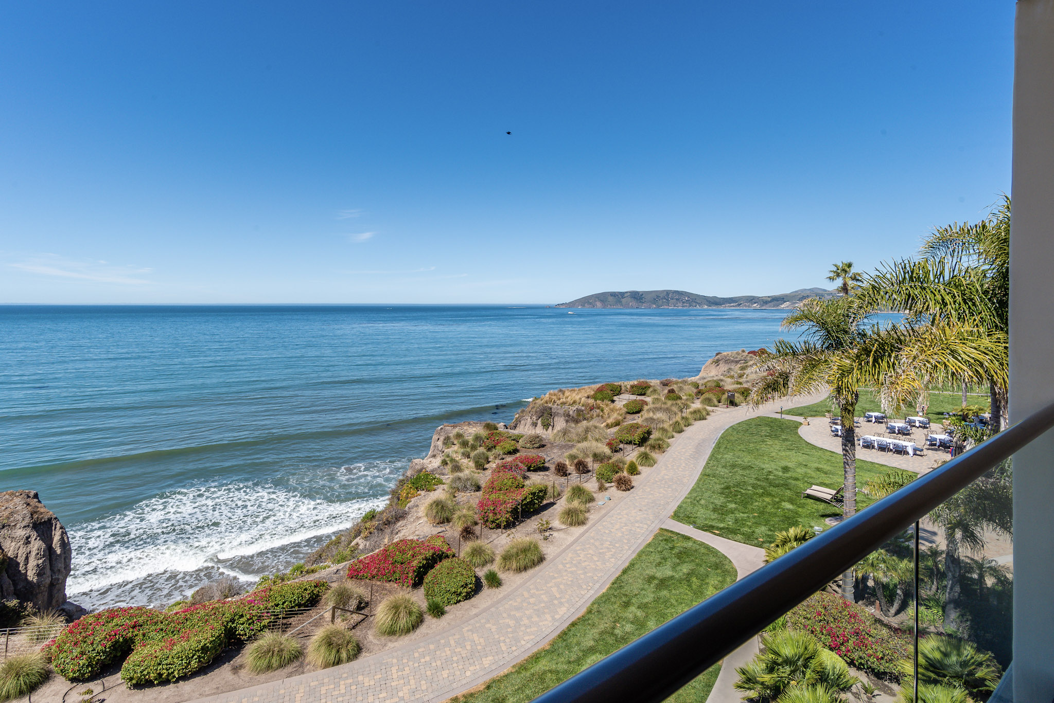Coastal scene with a sandy path, greenery, and ocean waves under a clear blue sky.