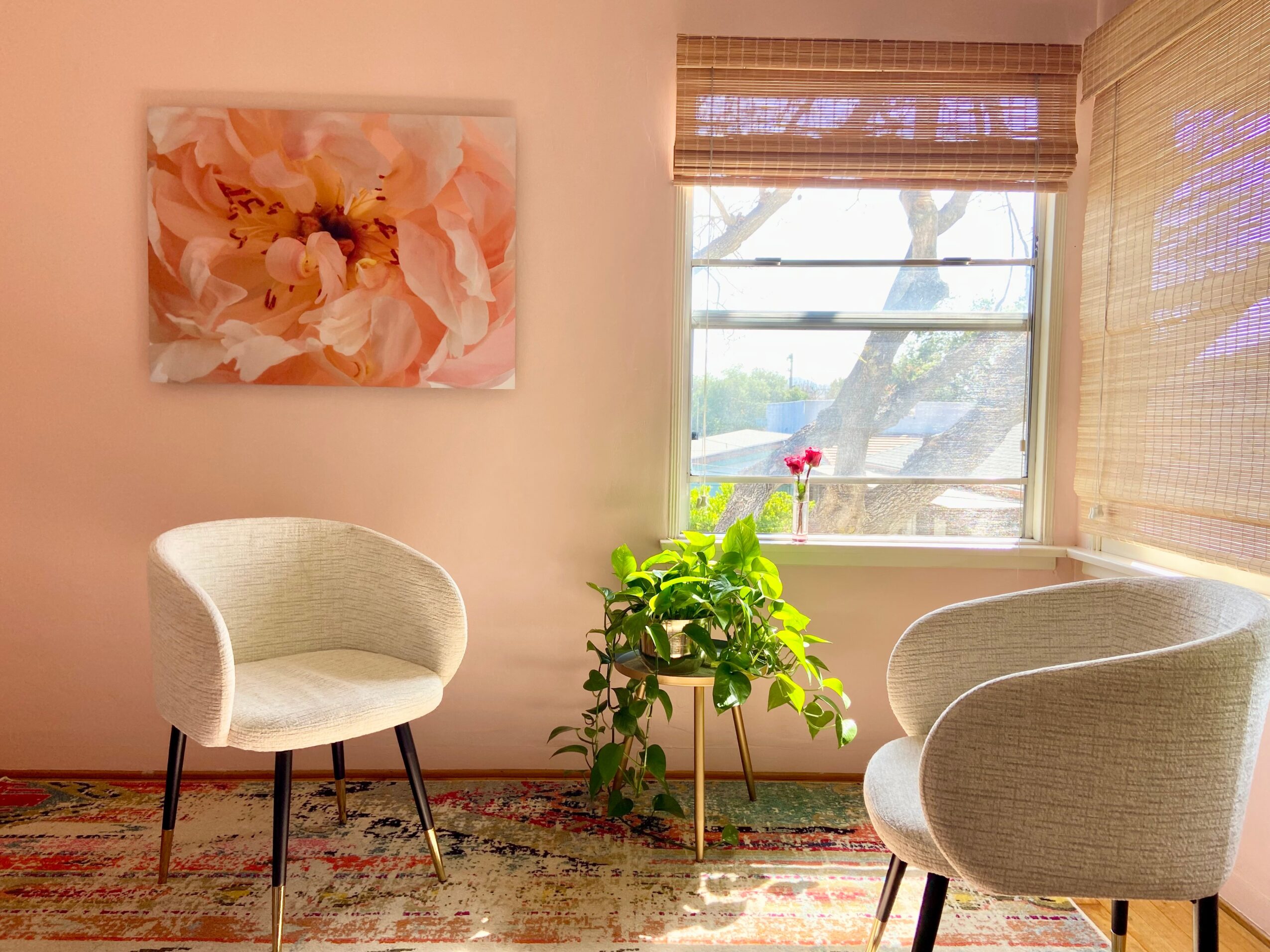 Two light-colored armchairs with dark legs are positioned near a window with blinds, a potted plant between them, and a floral painting on the wall.