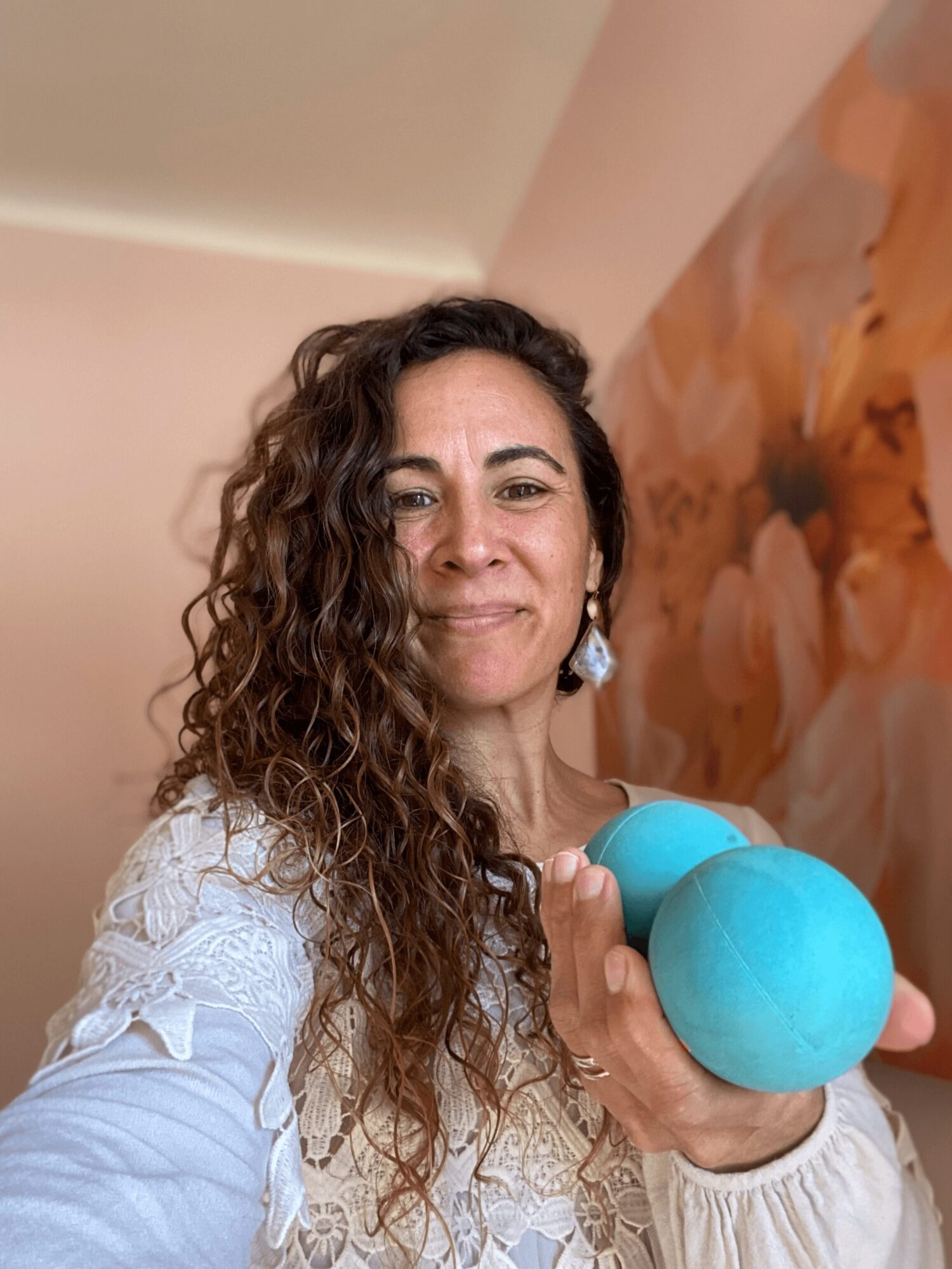 Woman with curly hair holding a blue dumbbell, smiling, in a room with a floral painting background.