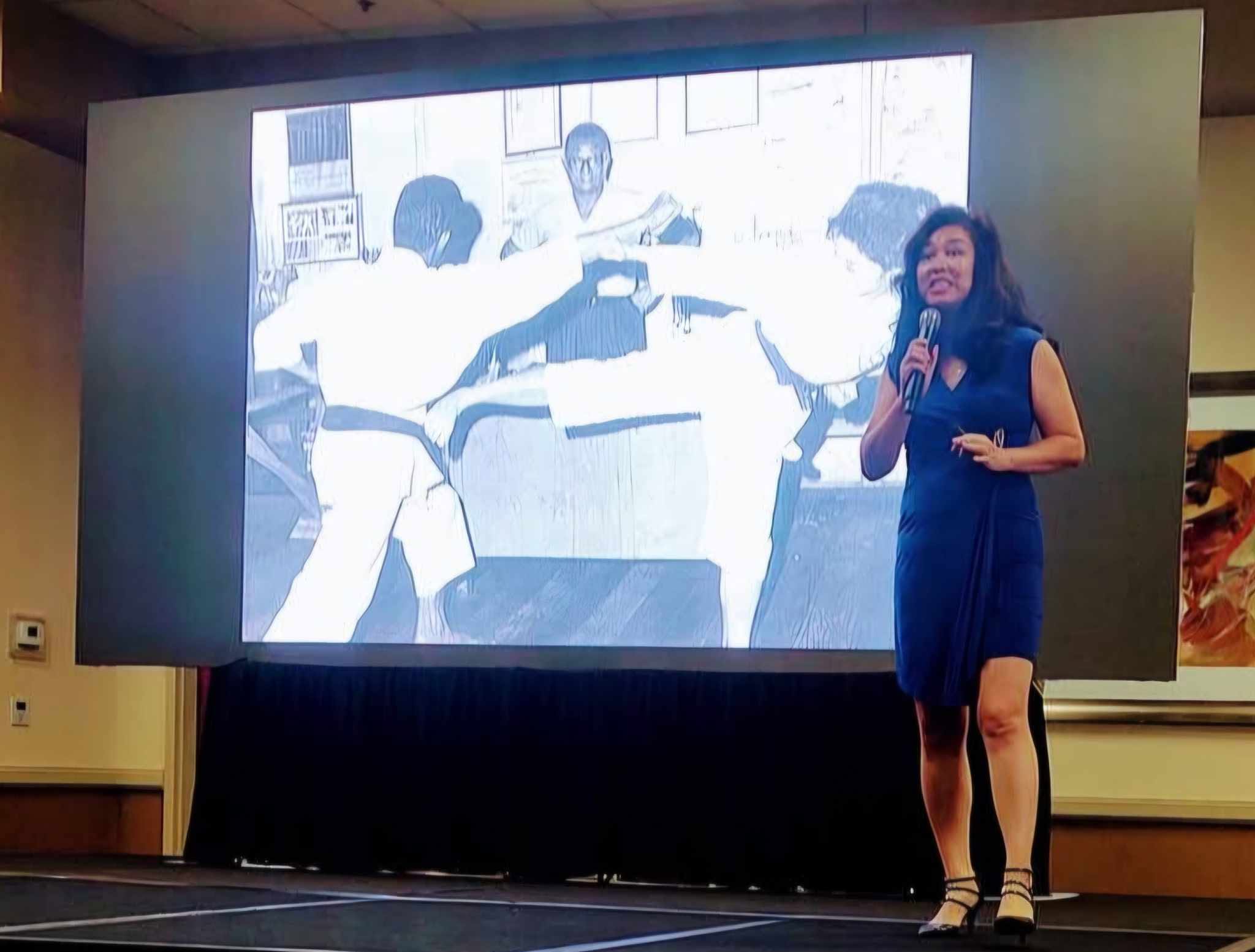 Woman in a blue dress holding a microphone, standing on stage in front of a large screen displaying a black-and-white photo of people practicing martial arts.
