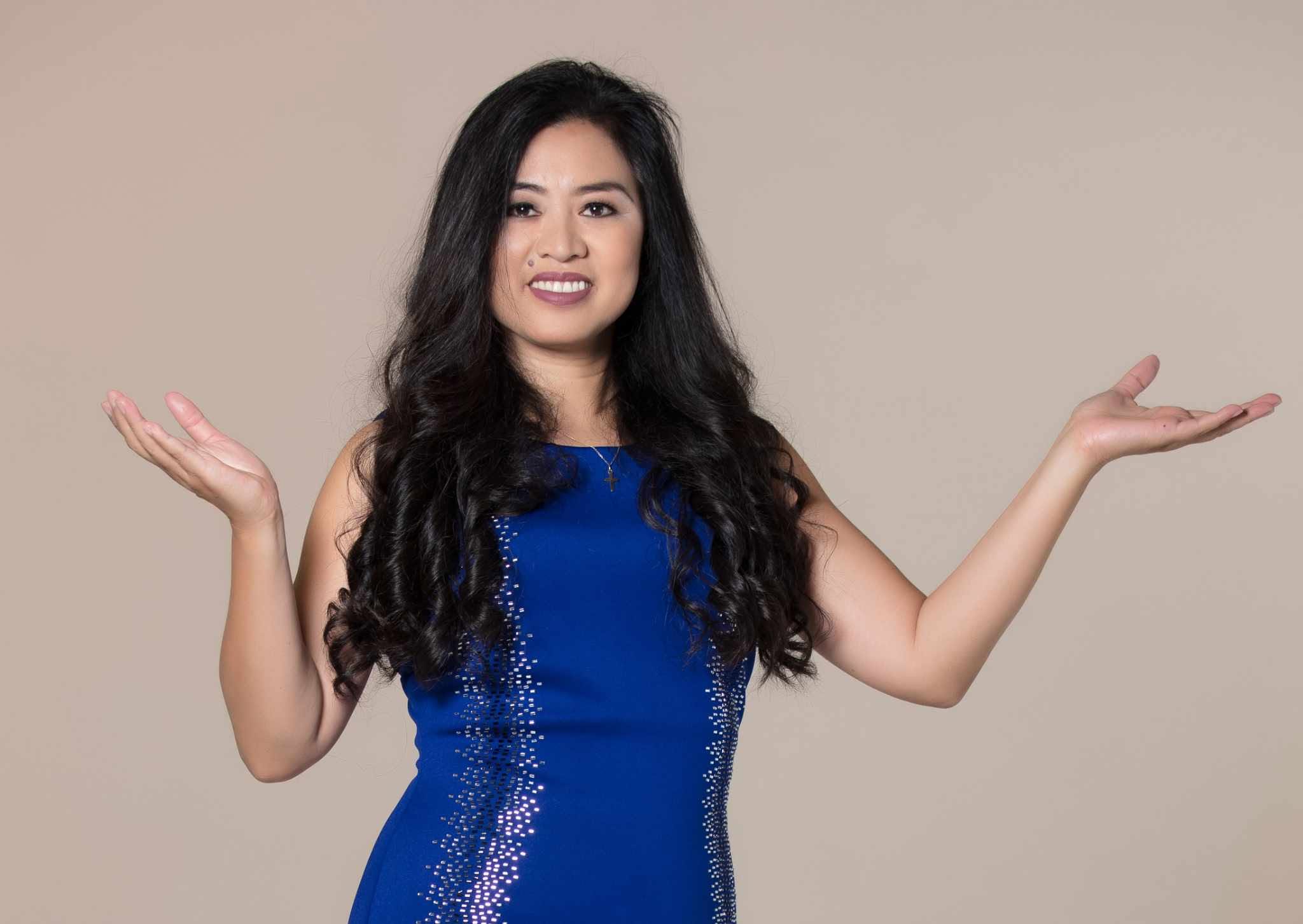 Woman with long dark hair wearing a blue dress, smiling, with arms raised and palms facing upward, against a plain background.