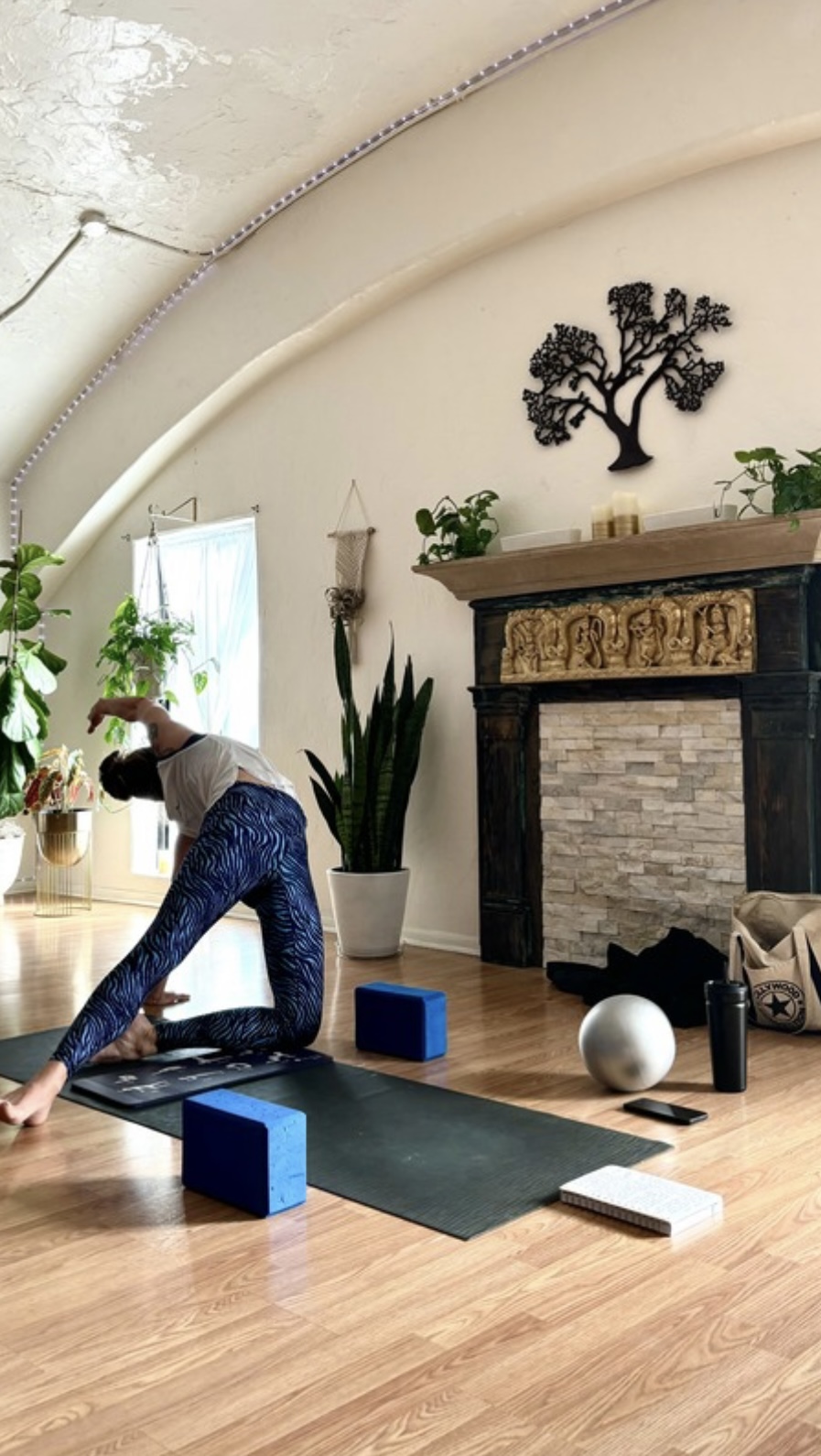 Person practicing yoga on a mat in a bright room with plants, a fireplace, and decorative wall art.