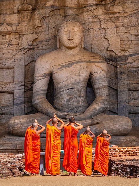 Five monks in orange robes sit in front of a large stone Buddha statue carved into a rock face.