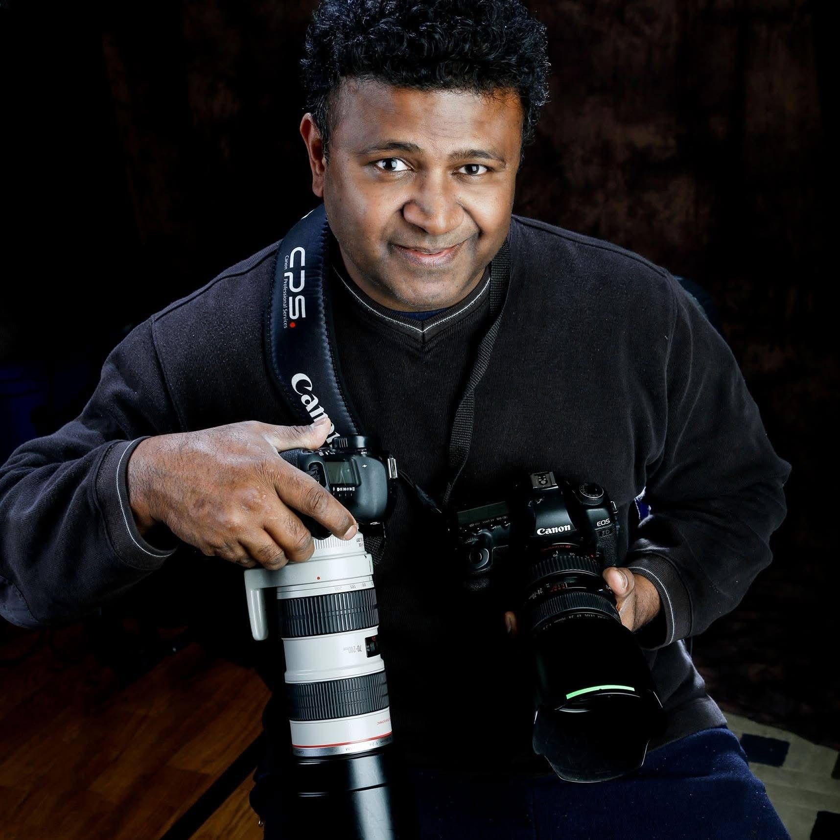 Man holding two cameras, one with a telephoto lens, smiling, dark background, wearing black jacket.