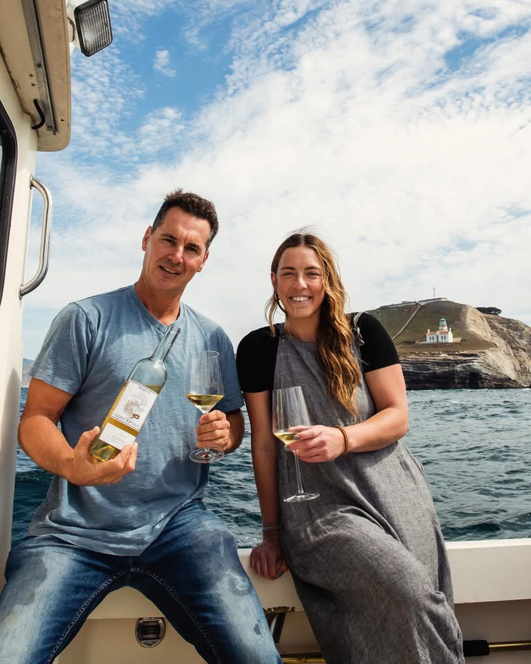 Two people smiling and holding wine glasses on a boat with water and a hill in the background.